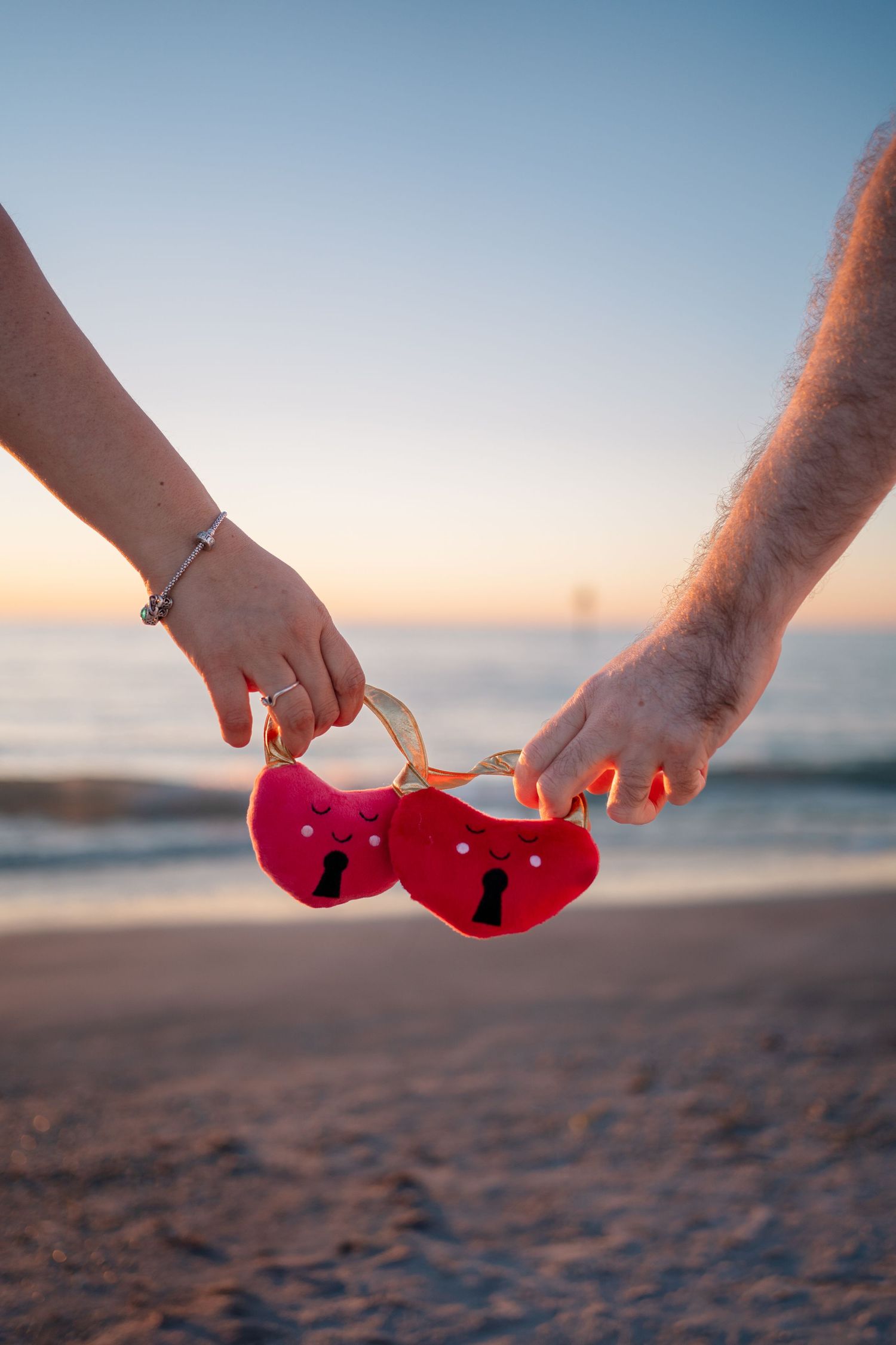 Couple holding red heart-shaped locks together on a sandy beach at sunset with ocean in the background.
