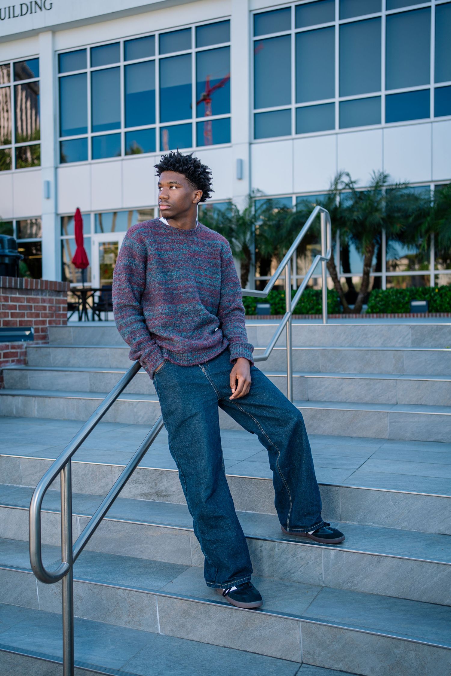 Young Black man in striped sweater and wide-leg jeans poses on outdoor steps near a modern building with palm trees.