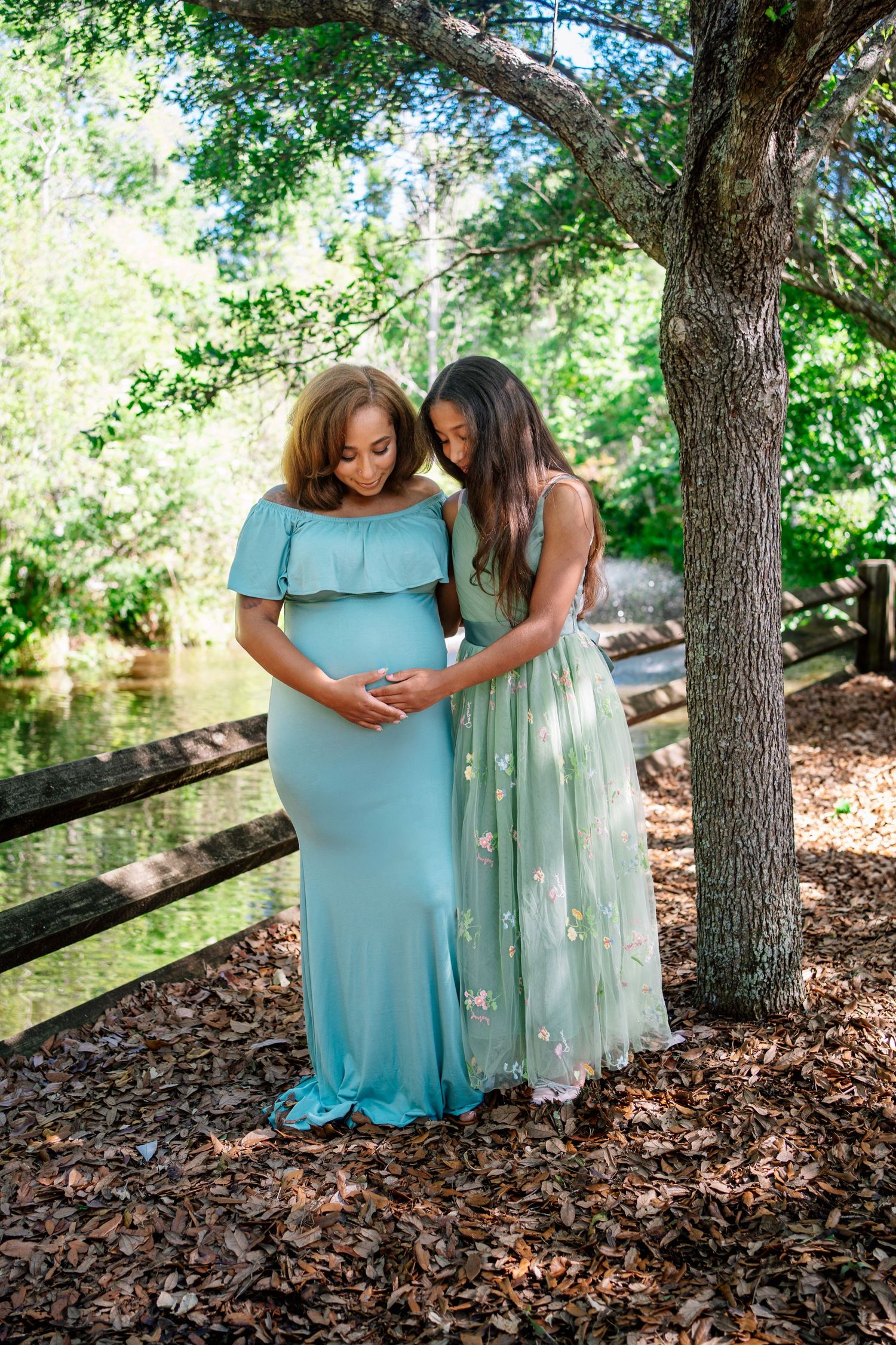 Two women in mint green gowns share a tender maternity moment outdoors near a wooden fence and pond.