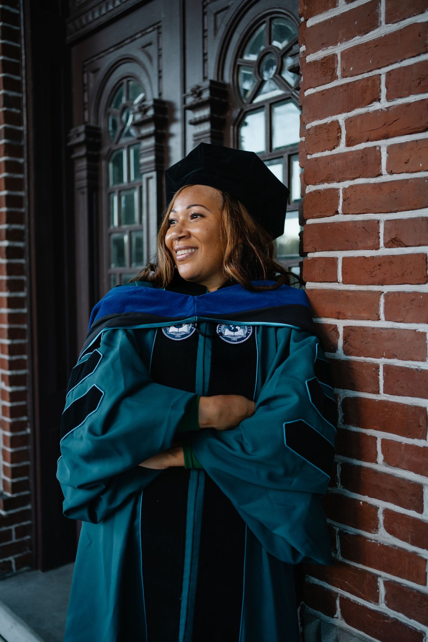 Smiling graduate in teal doctoral gown and blue hood leans against brick wall outside ornate wooden doors.