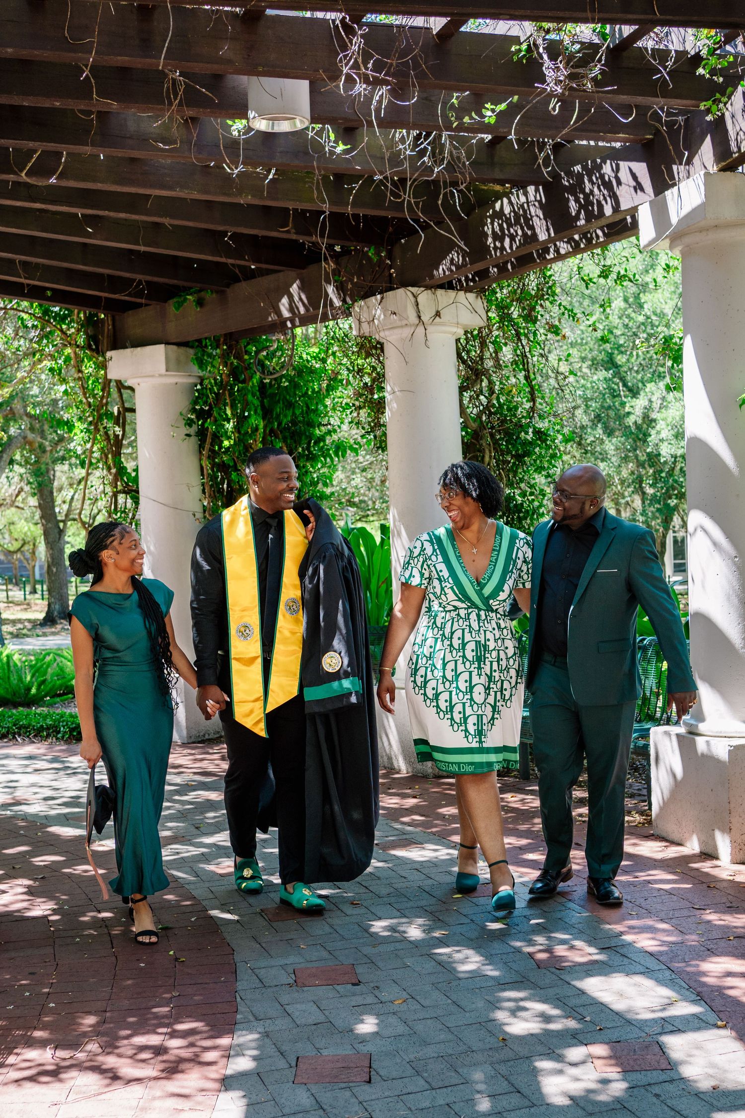 A graduate in cap and gown walks with three family members dressed in green attire on a brick path.