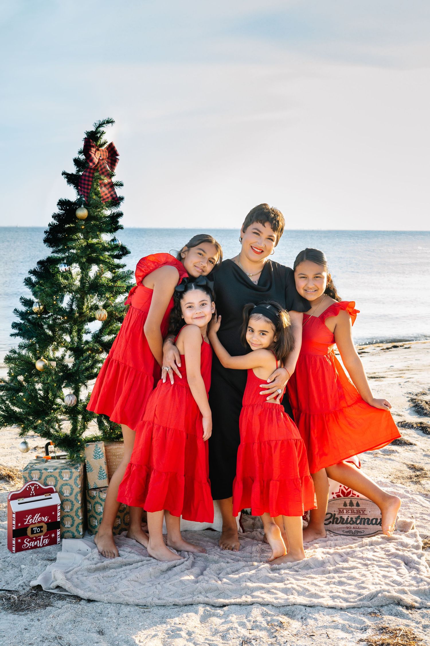 A mother and four daughters in red dresses pose by a Christmas tree on a sandy beach with ocean views.