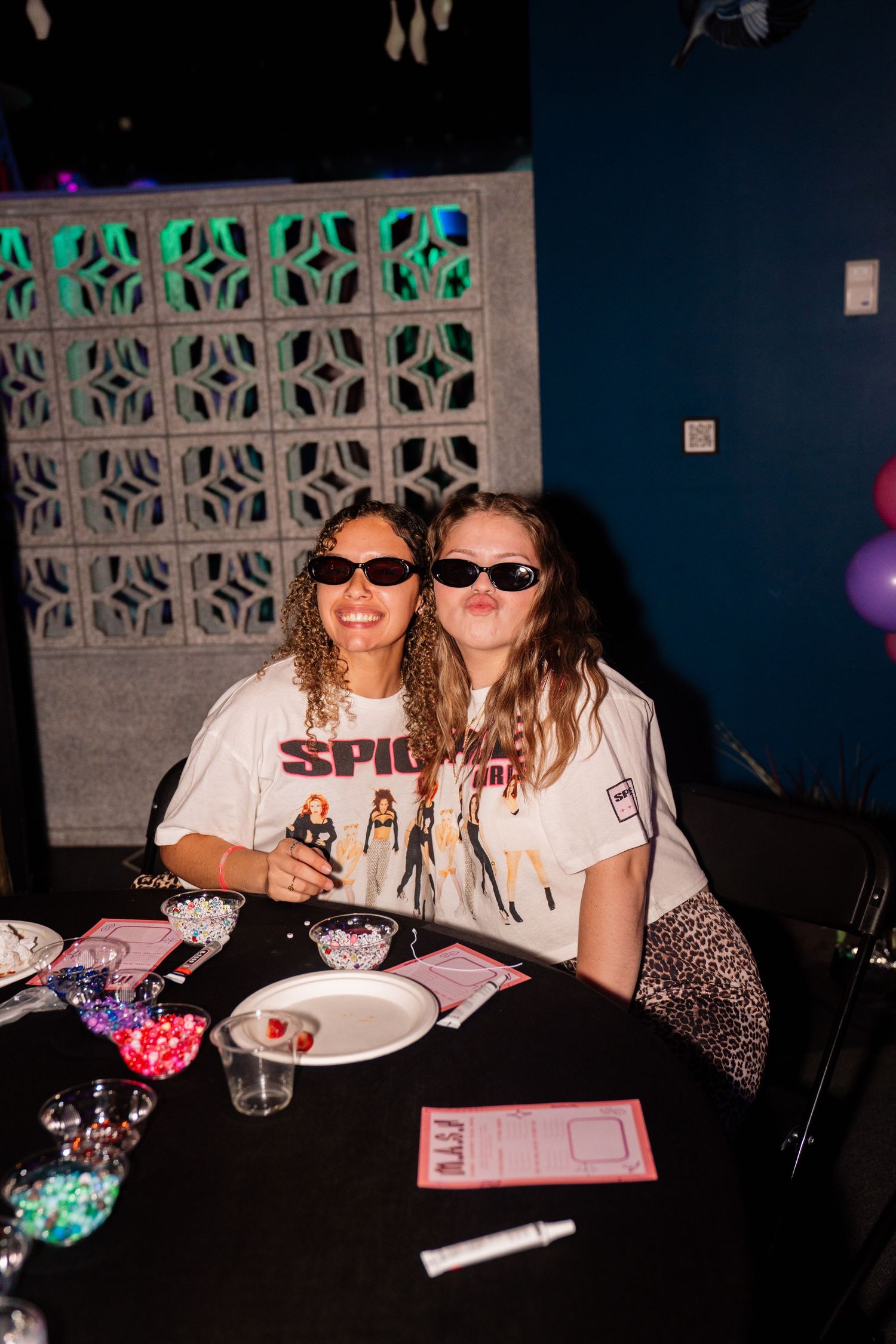 Two women in Spice Girls shirts and sunglasses smile at a party table with colorful decorations.