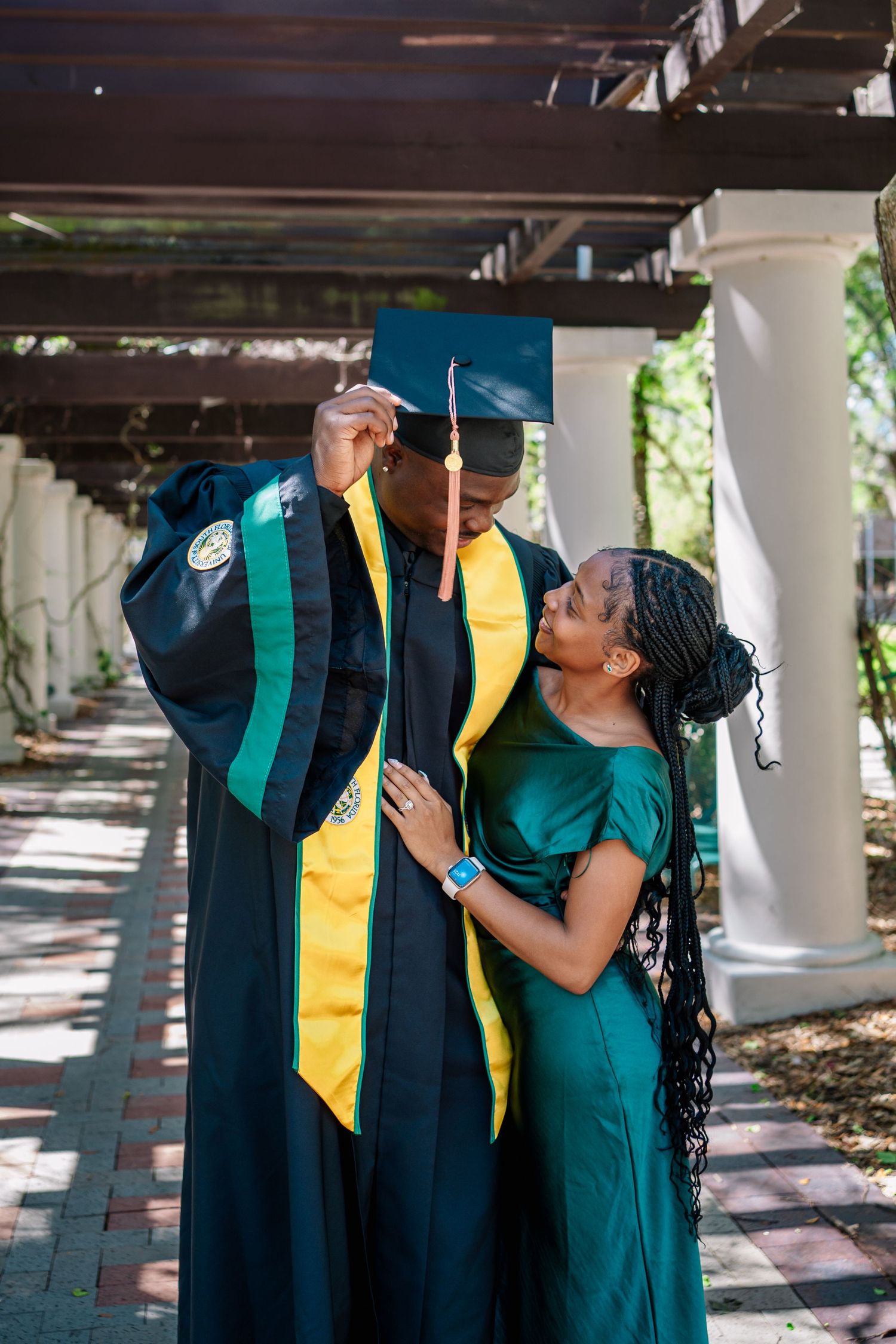 A graduate in cap and gown shares a tender moment with a woman in a green dress under a pergola.