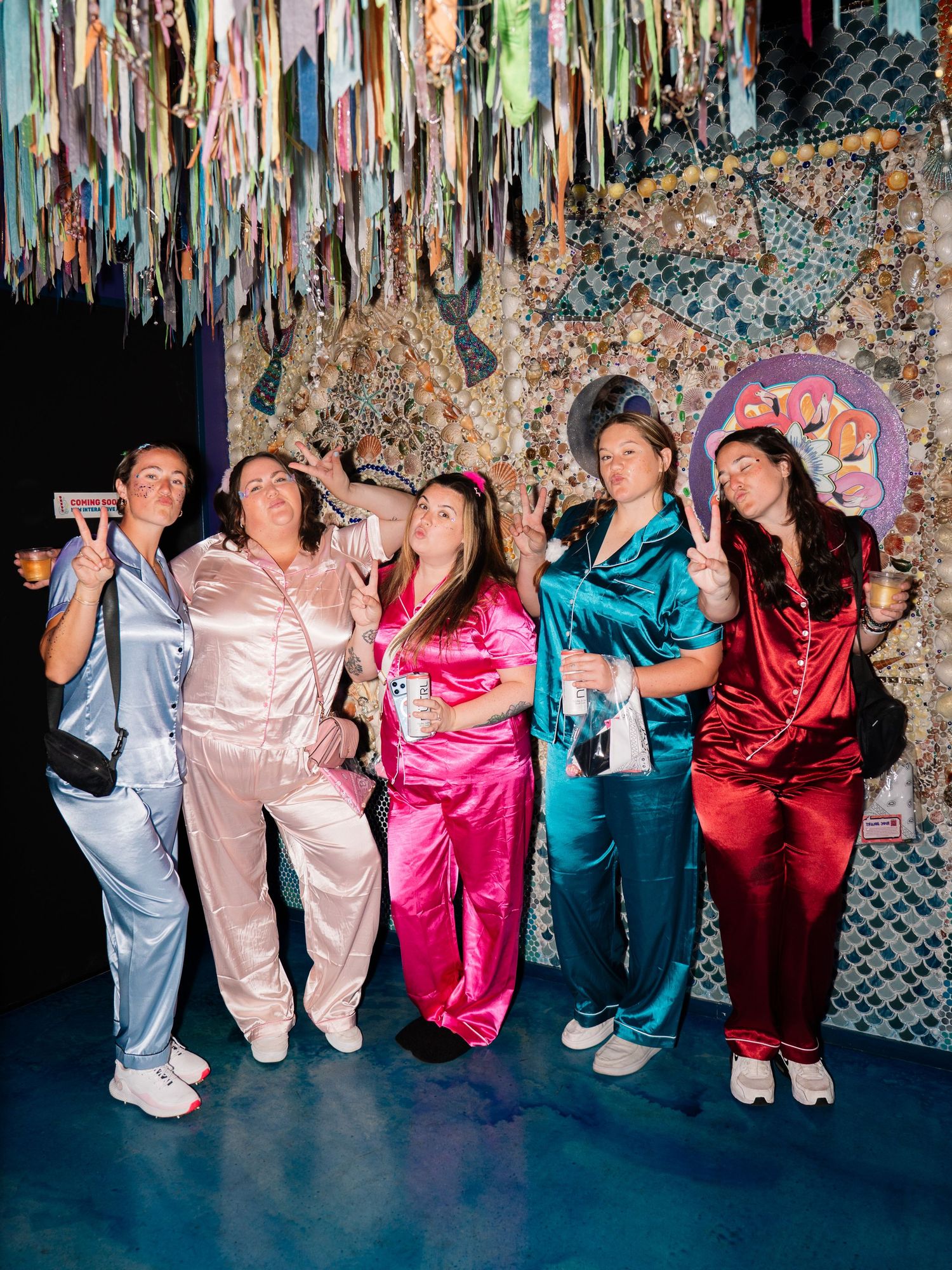 Five women in colorful satin pajamas pose together at a festive party with ribbon decorations overhead.