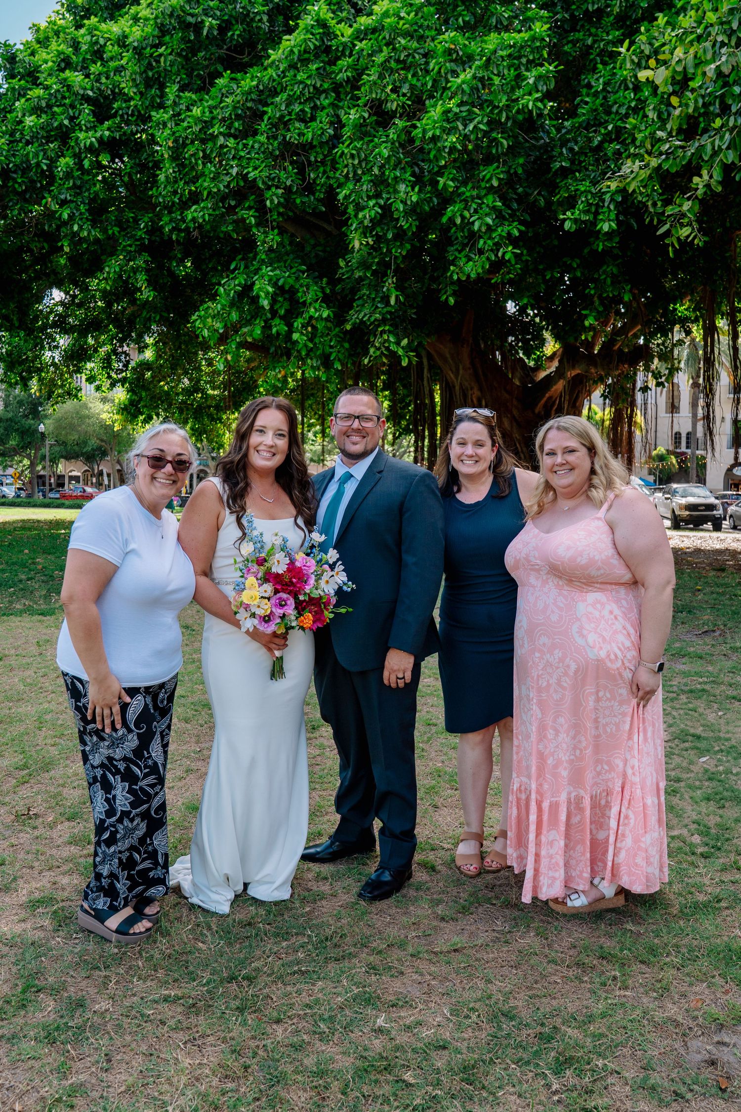 Wedding couple poses with three guests under a lush green tree, bride holding a bright bouquet in a white dress.