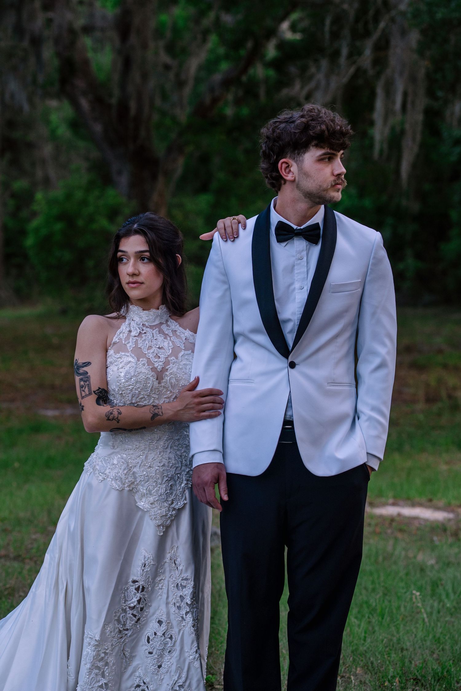 A bride in lace gown and tattooed groom in white tuxedo pose elegantly outdoors among lush green trees.