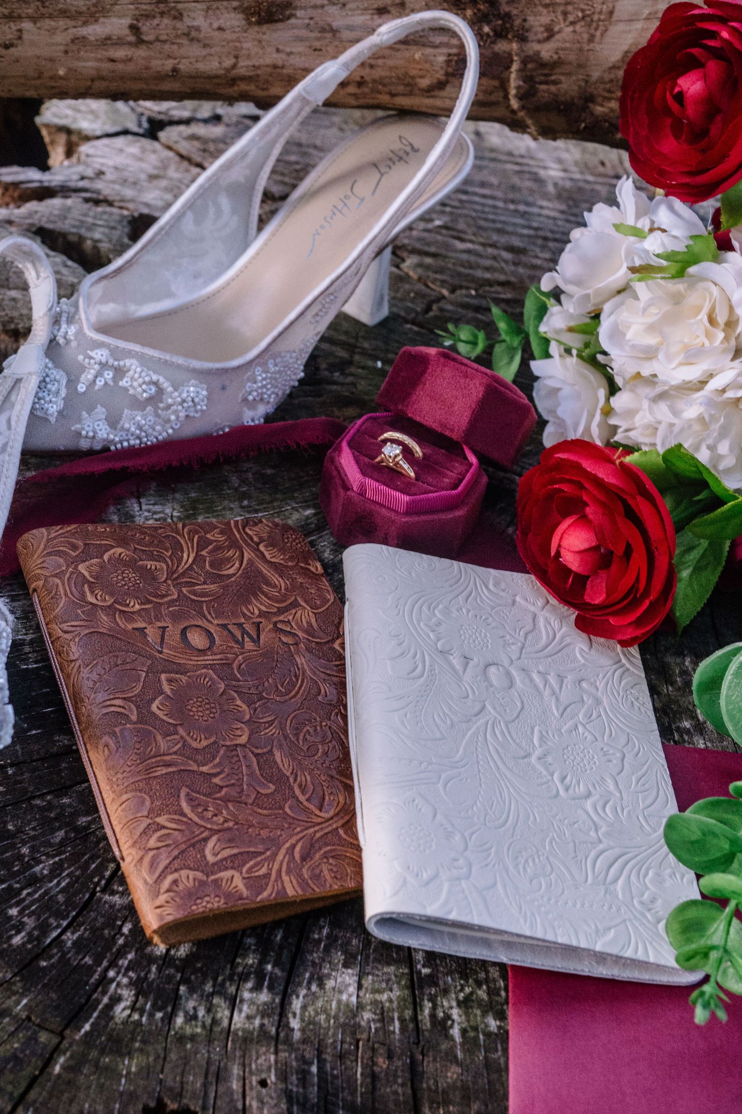 Wedding flat lay with vow books, diamond ring in velvet box, bridal heel, red and white roses on rustic wood.