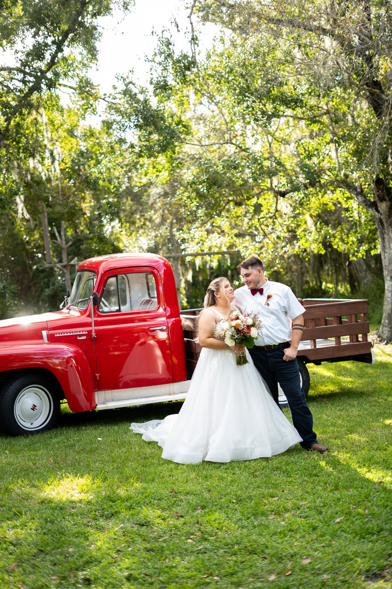 Romantic couple in wedding attire pose together on grass near a vintage red pickup truck in a sunlit garden setting.