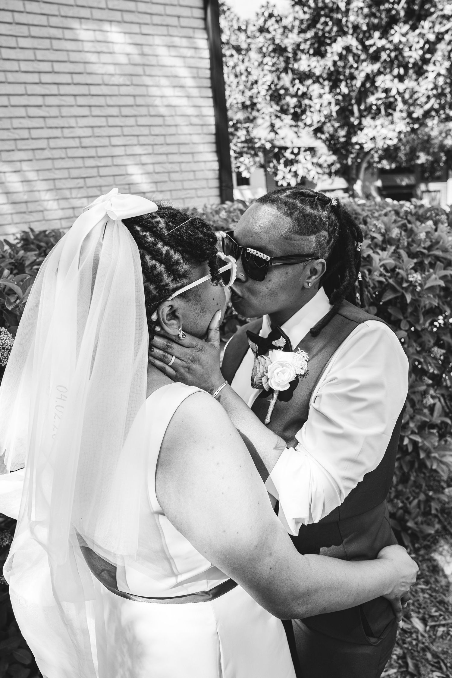 Black and white photo of a same-sex couple sharing a kiss on their wedding day, bride in veil and groom in vest.