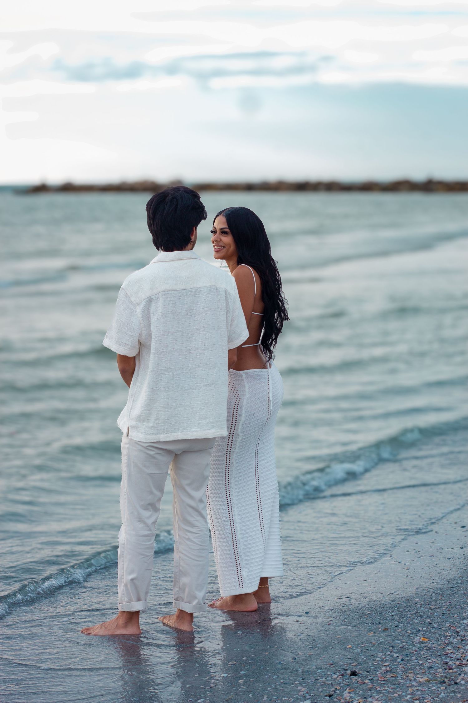 Couple walking along shoreline in white outfits during sunset photoshoot.