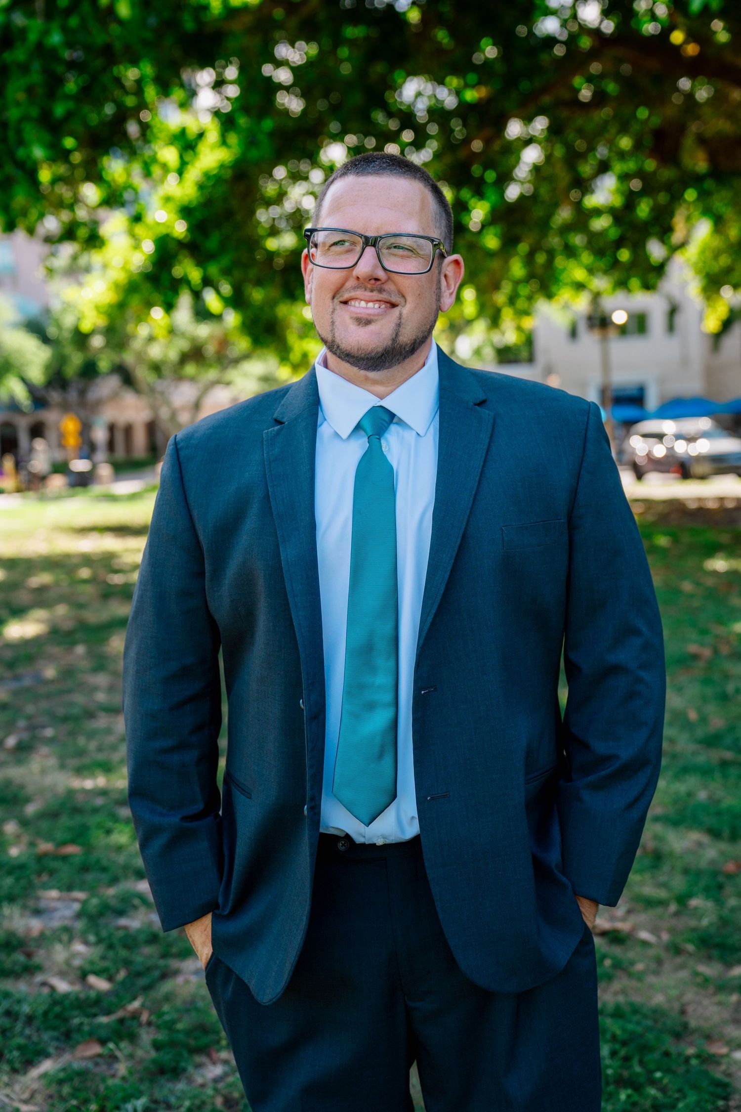 Smiling man in dark suit and teal tie wearing glasses stands outdoors under green trees.