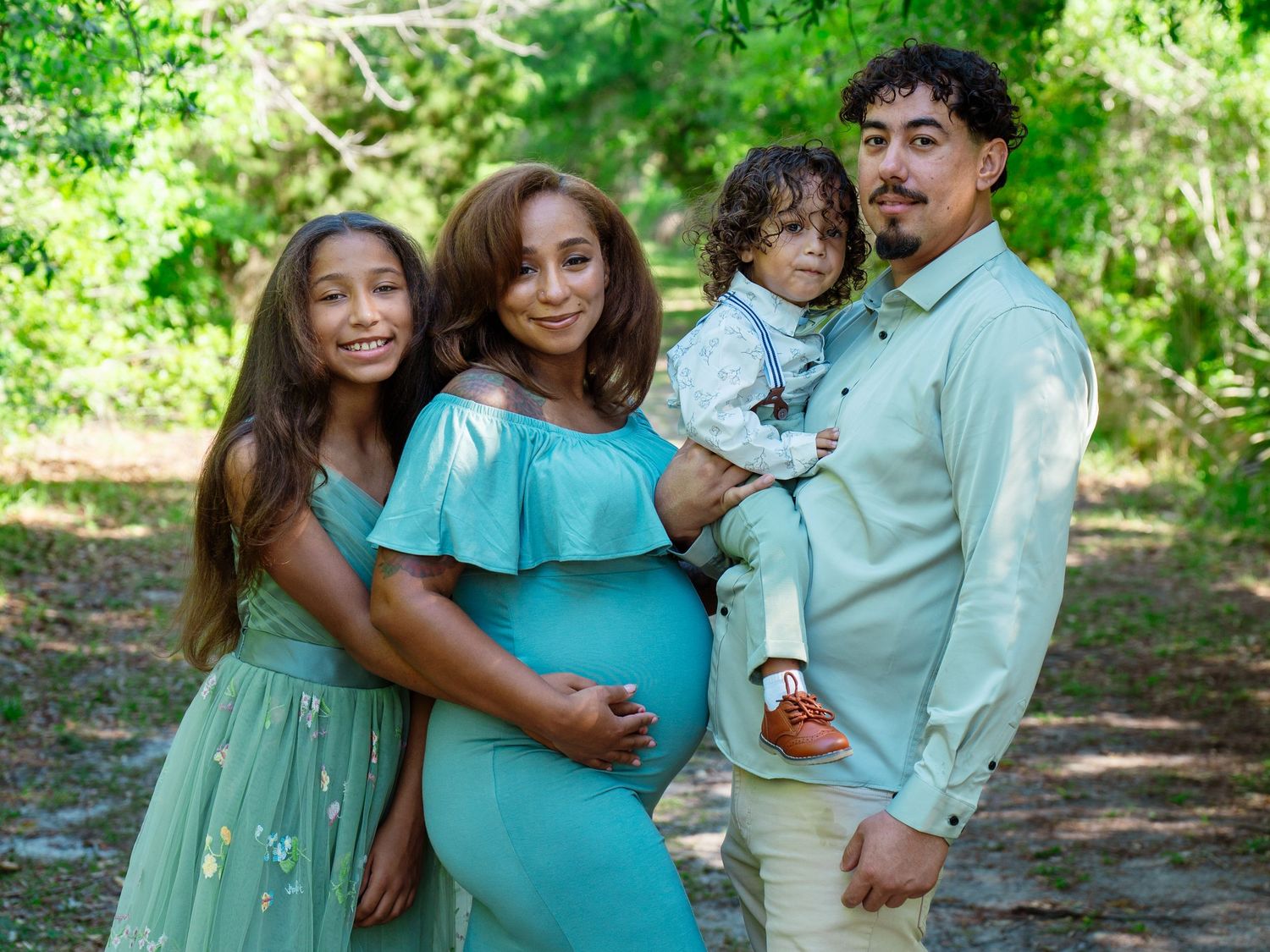 A pregnant woman in a teal dress poses outdoors with her family, including a daughter and husband holding a toddler.