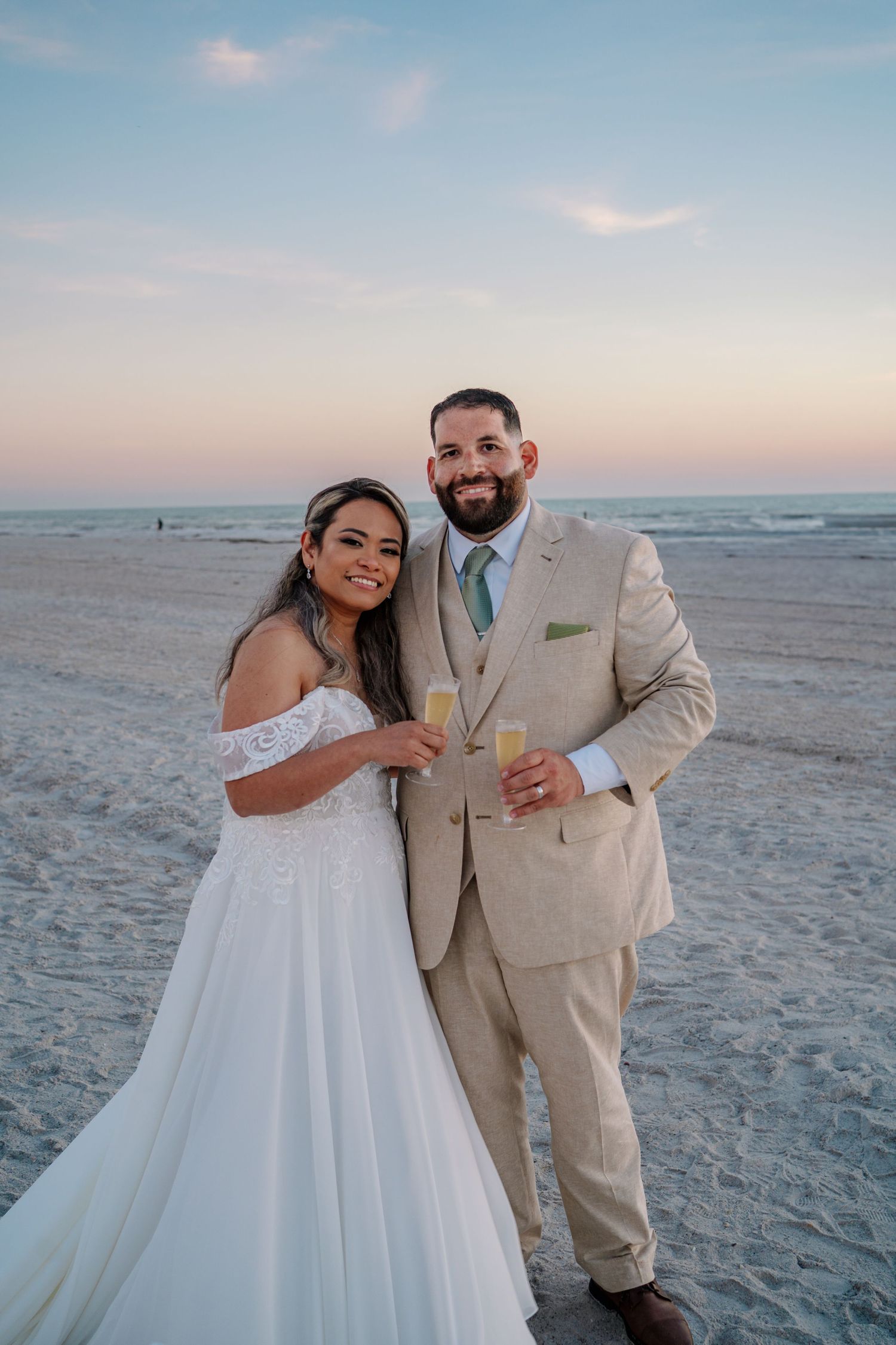 A couple in wedding attire holding champagne glasses poses together on a beach at sunset.