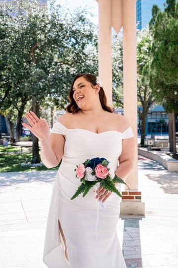 A bride in a white off-shoulder wedding dress poses outdoors with a pink and green bouquet under a covered walkway.