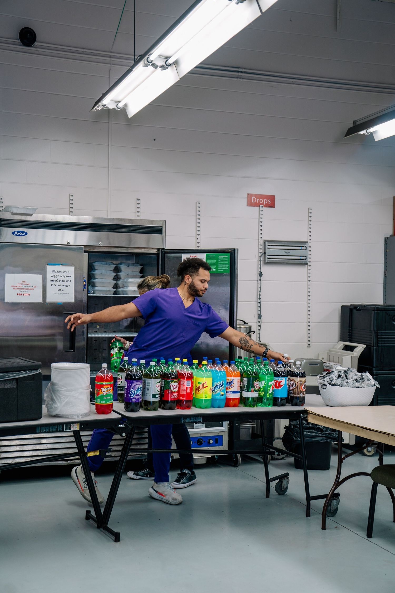 Man in purple uniform arranging colorful soda bottles on a table in a commercial kitchen.