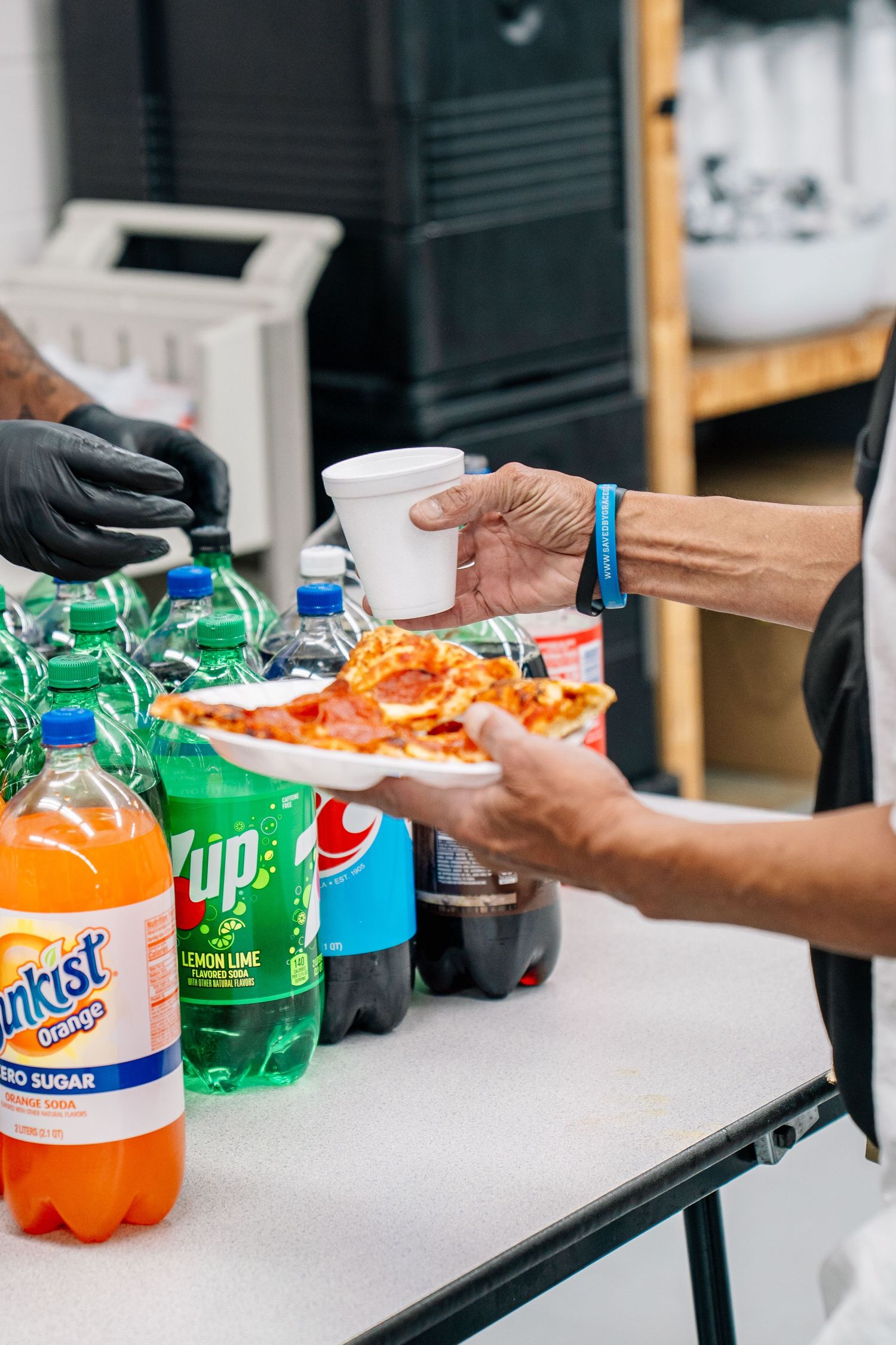 Person serving pizza slices and drinks including Sunkist, 7UP, and cola bottles at a food service table.