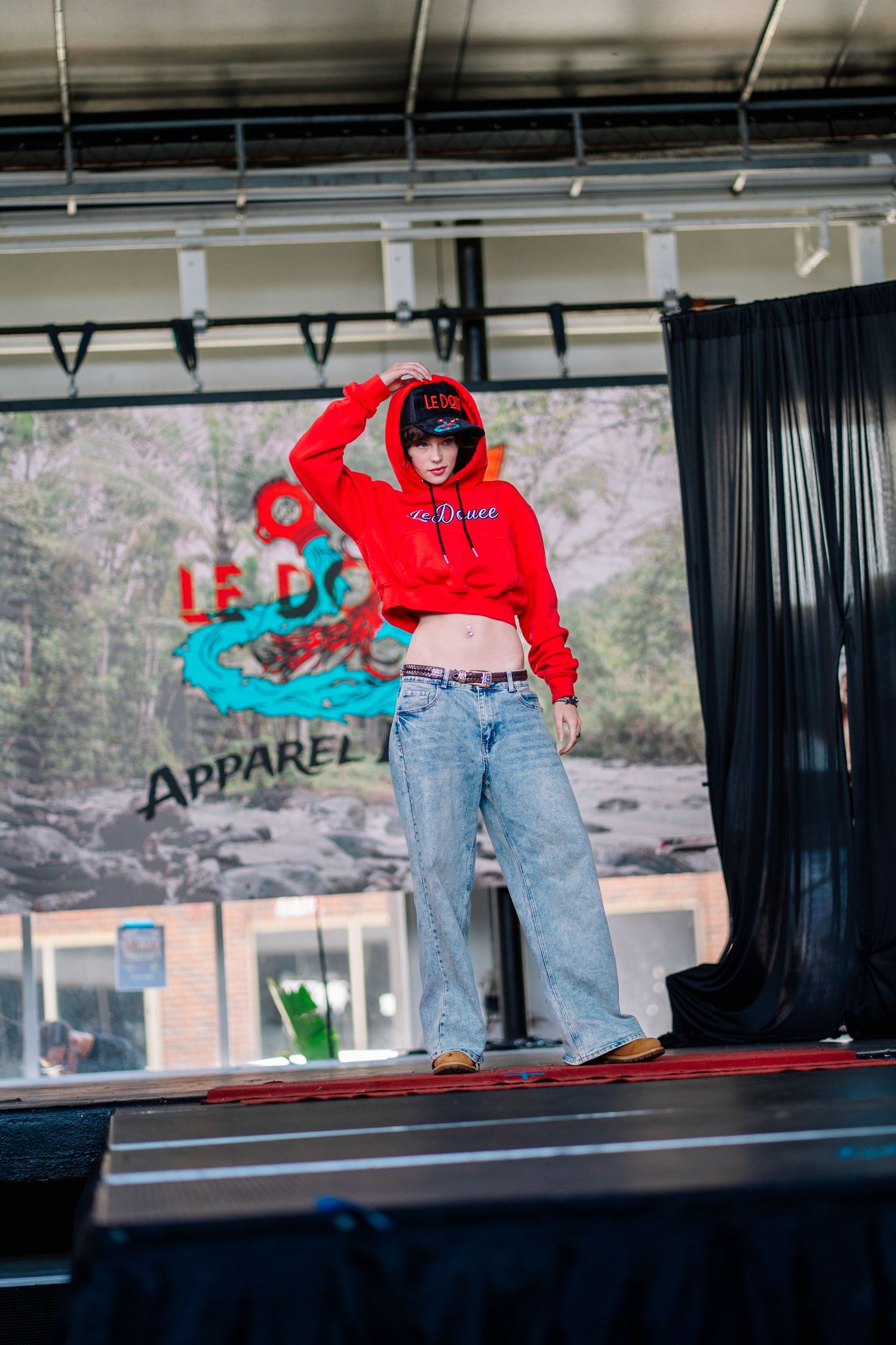 Model wearing red cropped hoodie and wide-leg jeans on a fashion runway stage.