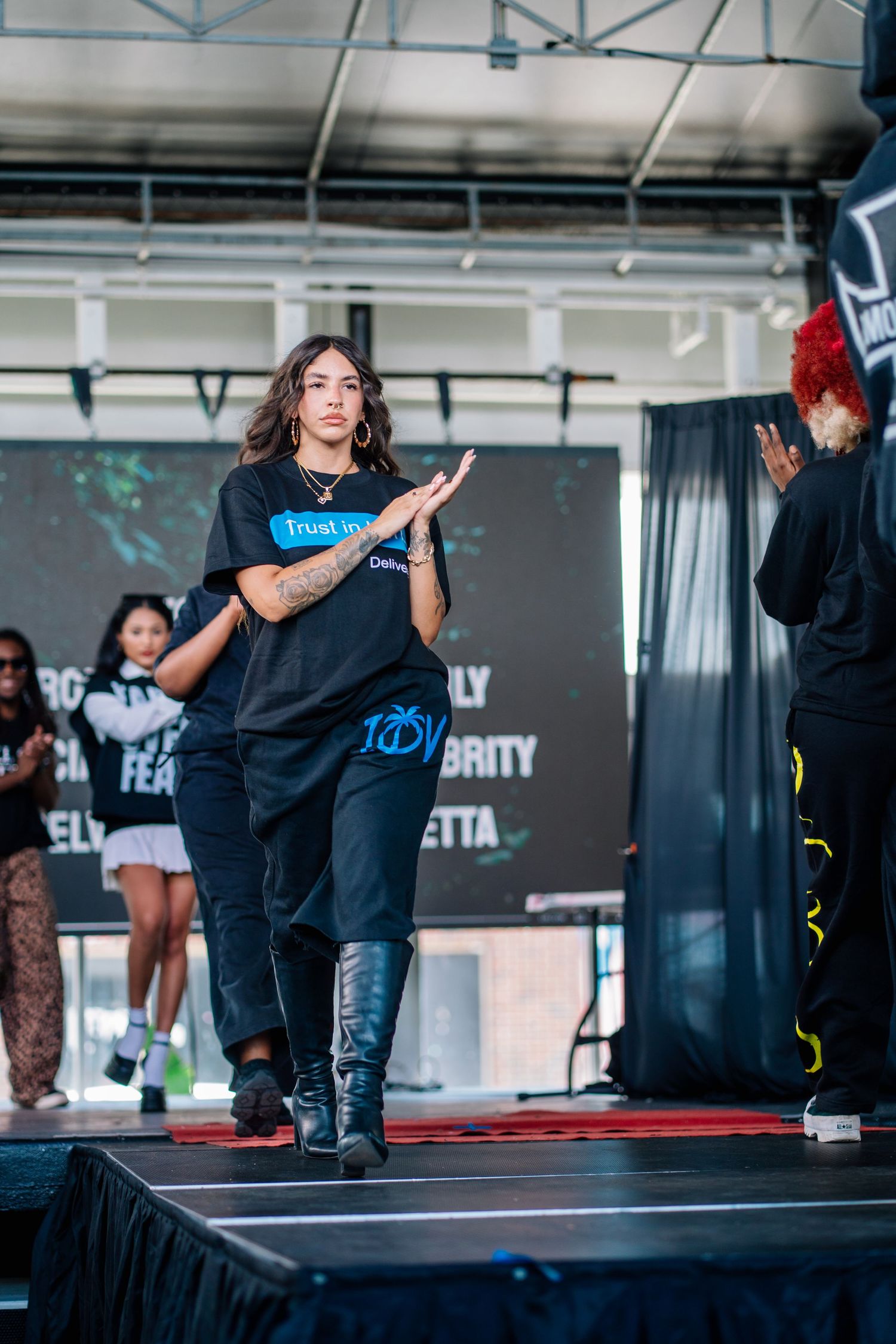 Person in black t-shirt and boots clapping on stage at an outdoor event with performers in the background.
