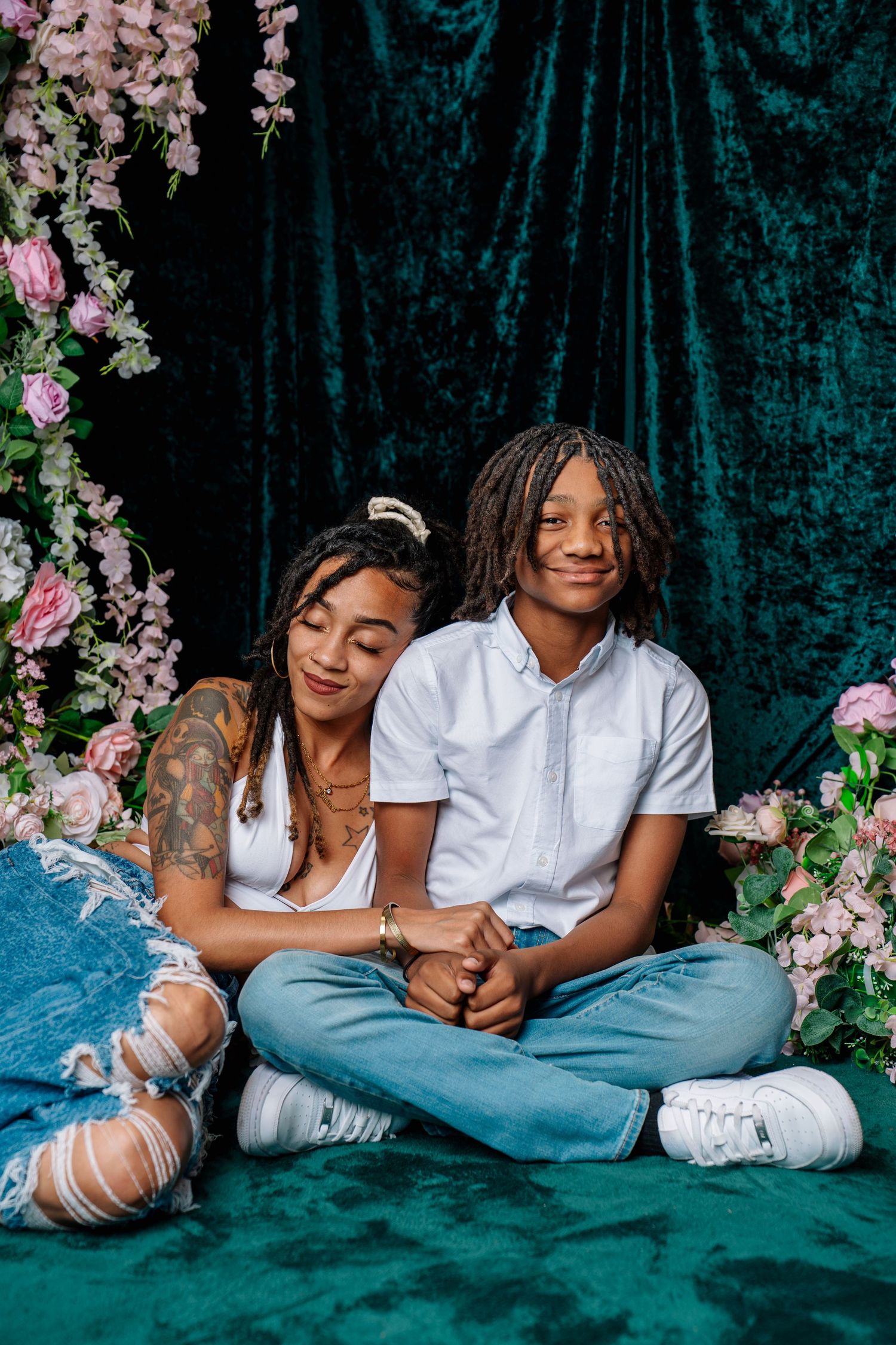 Two people sitting cross-legged in front of teal velvet backdrop with pink floral decor, wearing white tops and jeans.