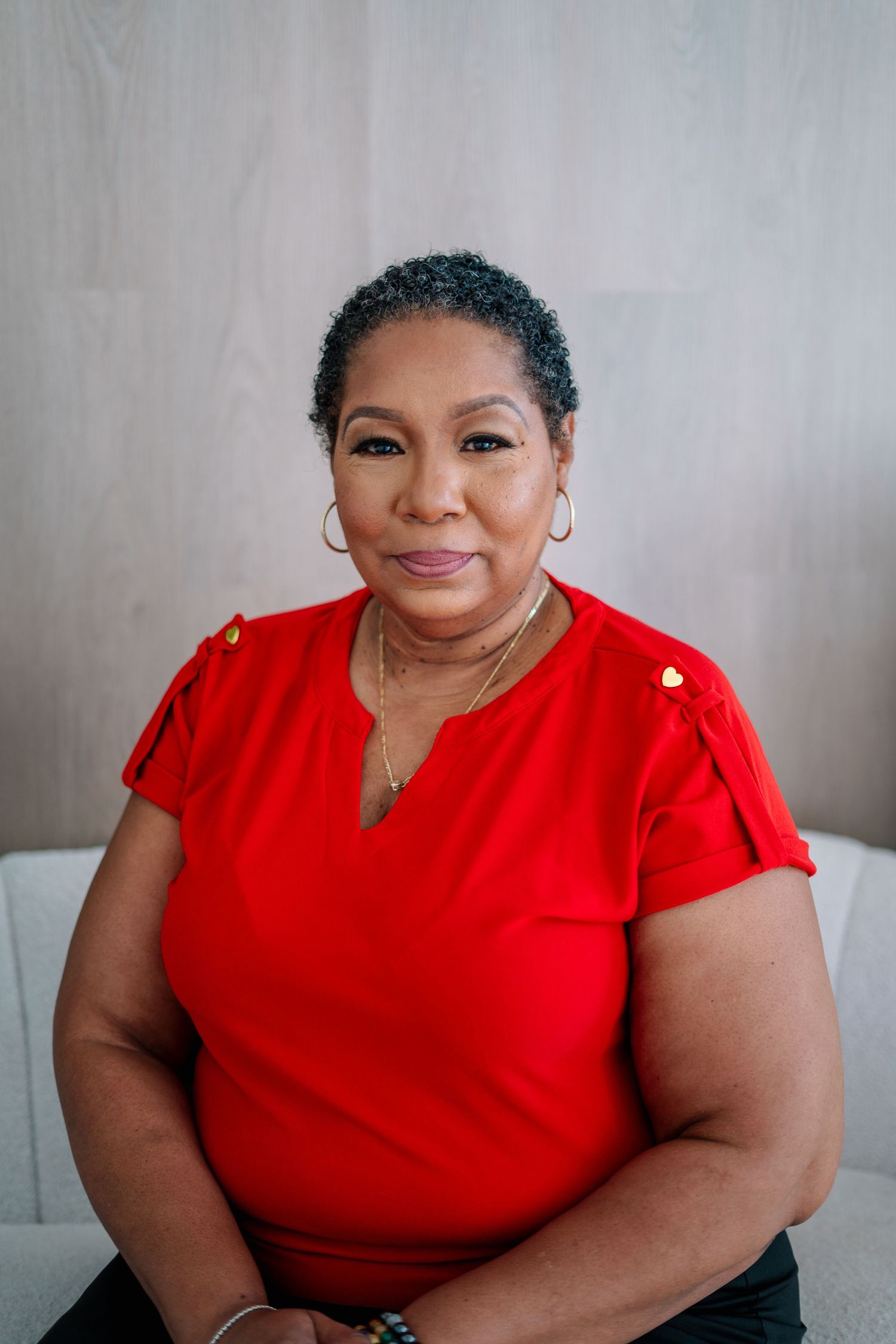 Middle-aged Black woman in red blouse with gold earrings and necklace sitting on white sofa against wood panel wall.