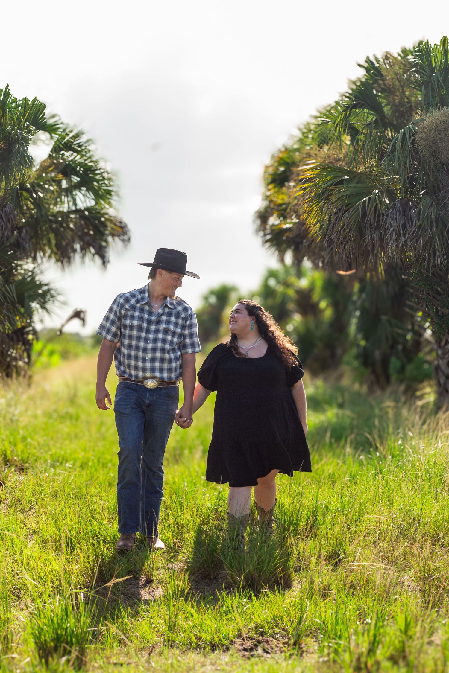 A couple walks through a lush field with palm trees on a cloudy day.