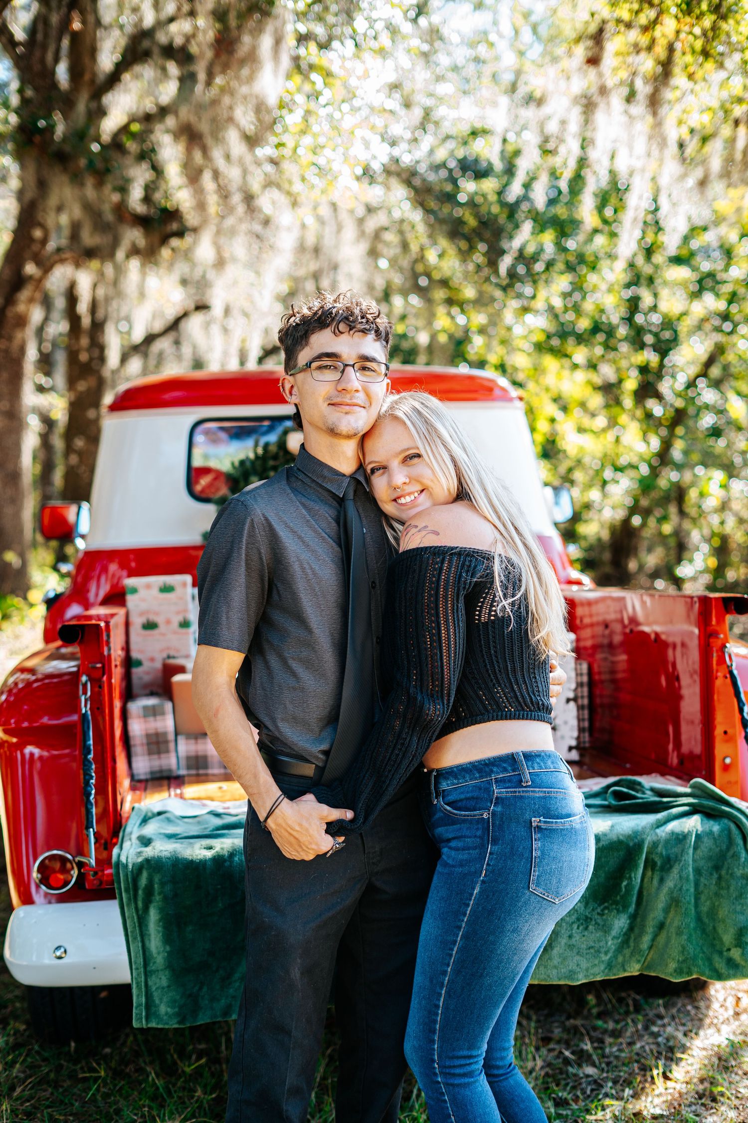 A couple shares tender moments in front of a vintage red truck on a sunny day in nature.