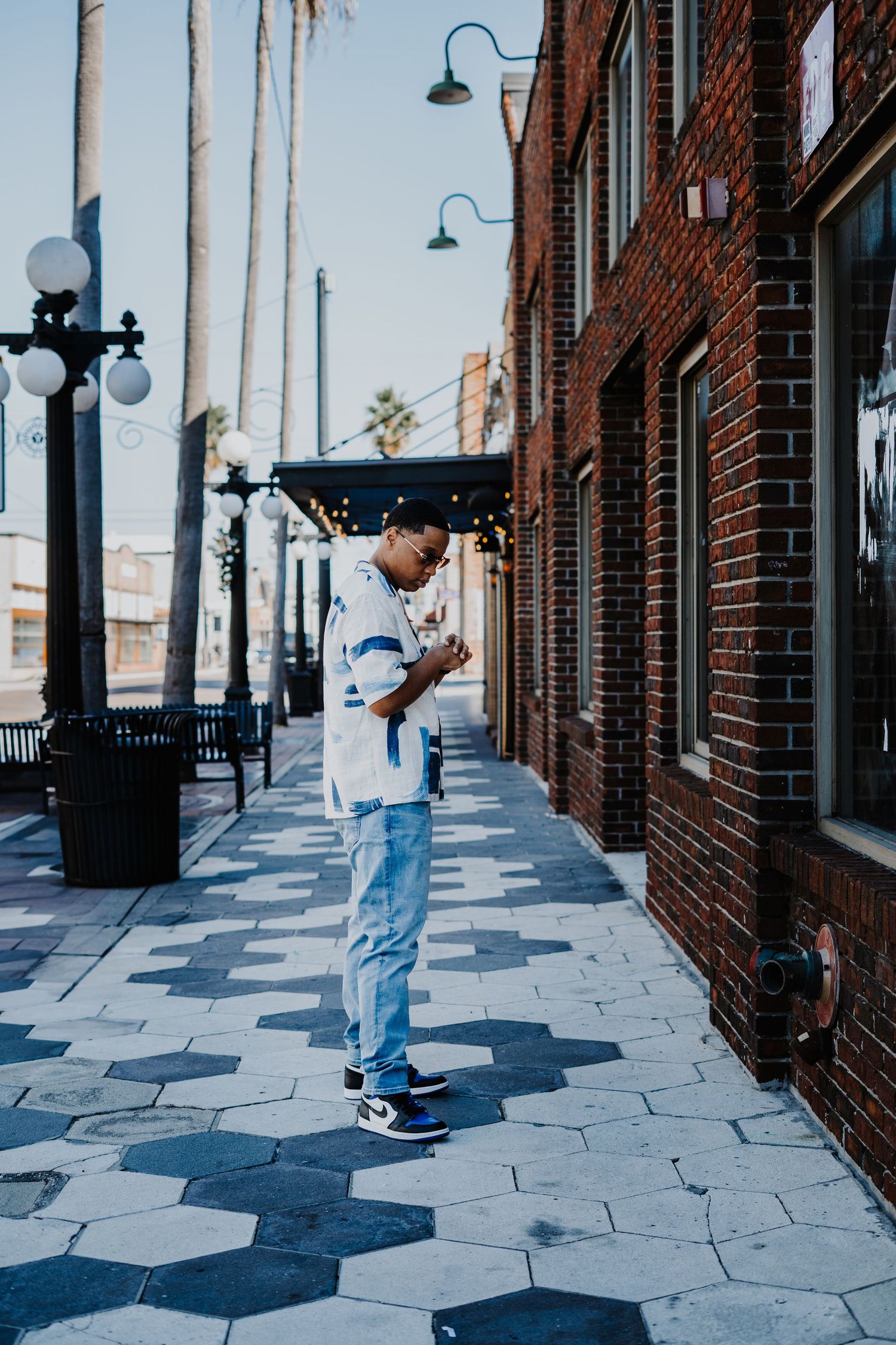 A person in light blue clothing performs skateboarding tricks on a patterned sidewalk next to brick buildings.