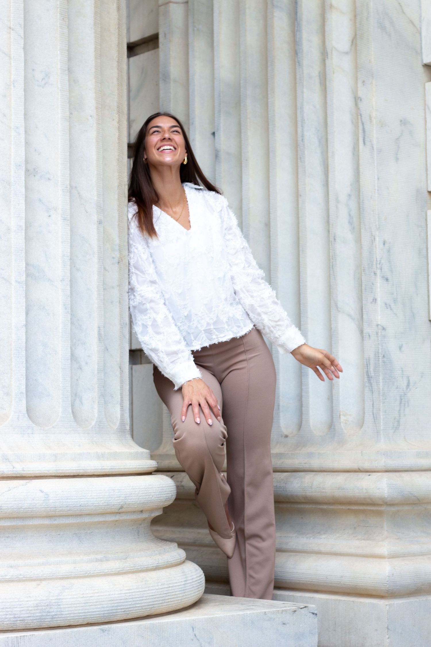 A person in a white blouse and brown pants poses playfully against classical stone columns in a photo series.