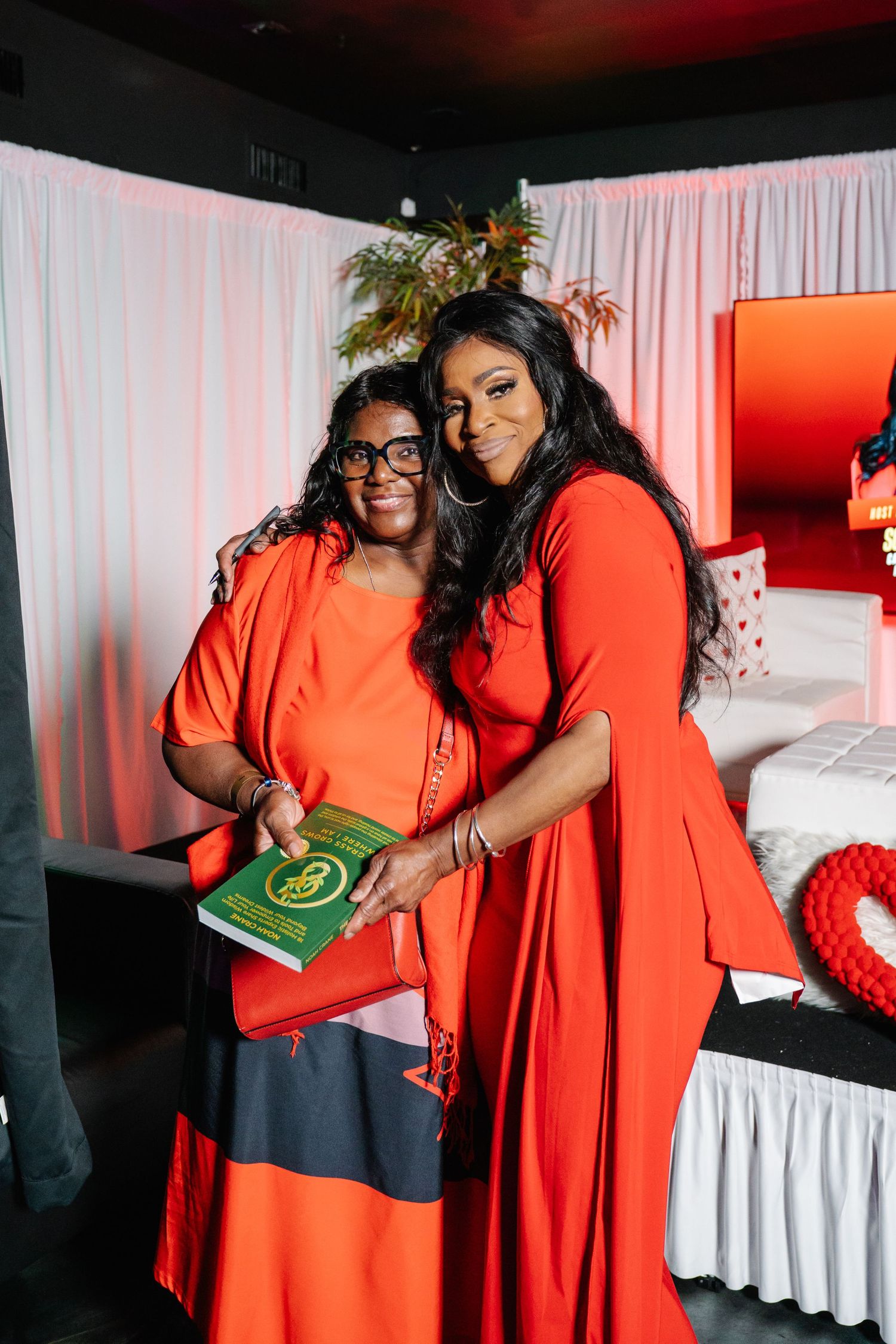Two women in matching red outfits pose together at an event, one holding a green book.