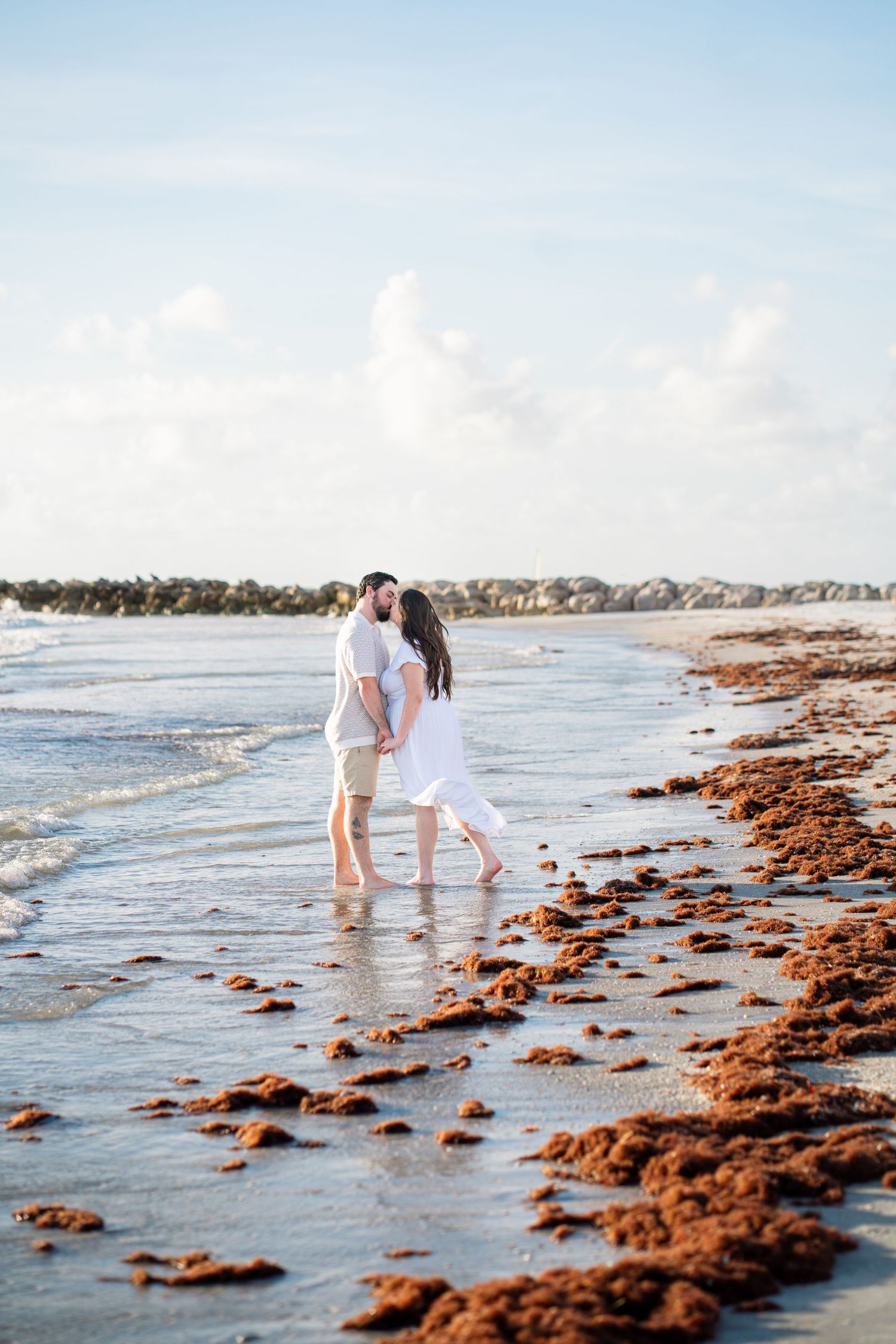 Couple sharing a romantic kiss on a sandy beach with red seaweed shoreline and calm ocean waves behind them.