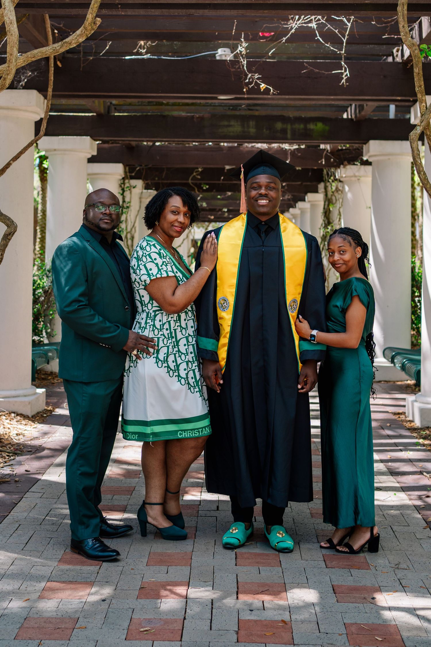 Family celebrates college graduation outdoors, graduate in black cap and gown with gold stole, surrounded by loved ones in green attire.