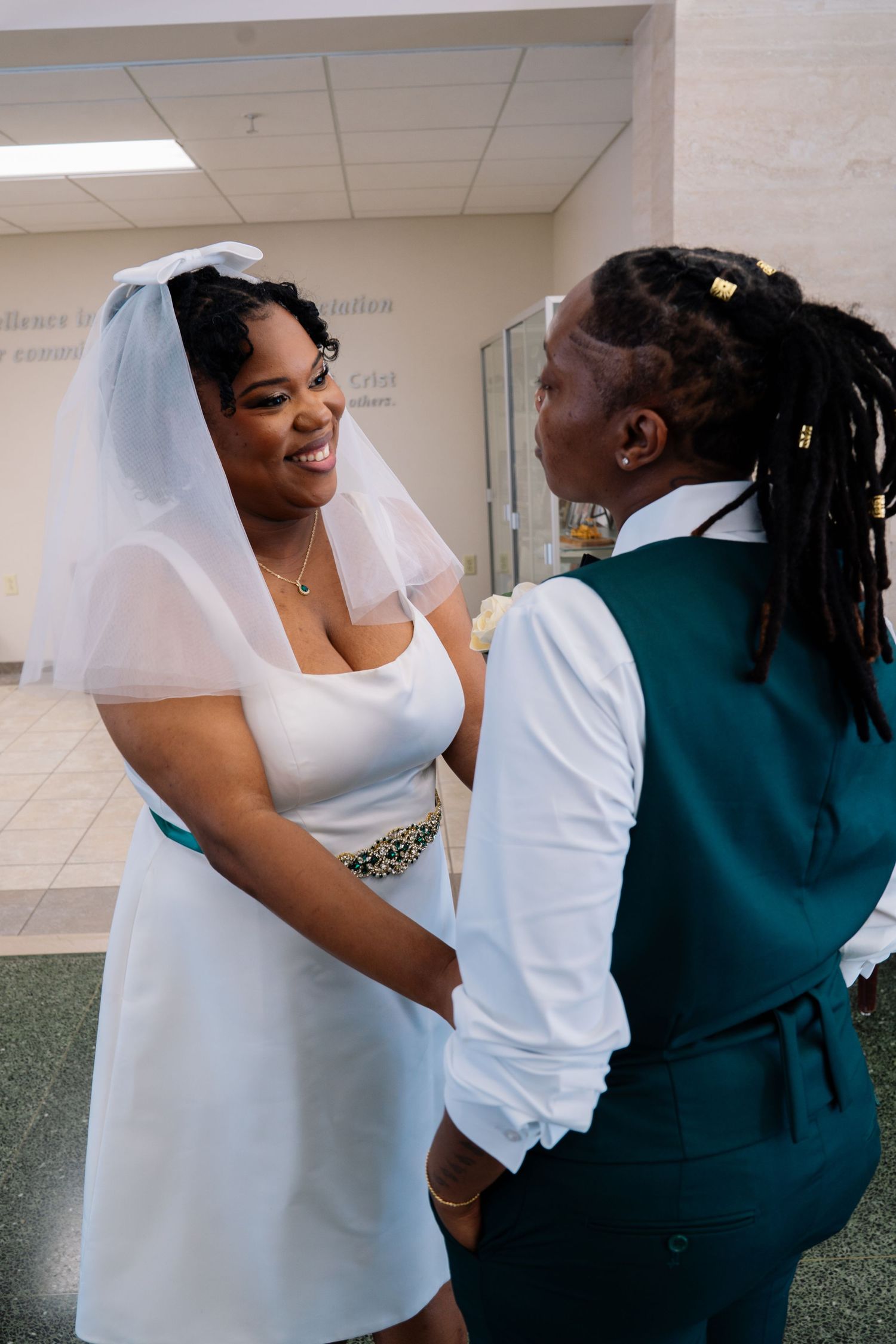Two women celebrate their wedding, one in a white dress and veil, the other in a teal vest, holding hands.