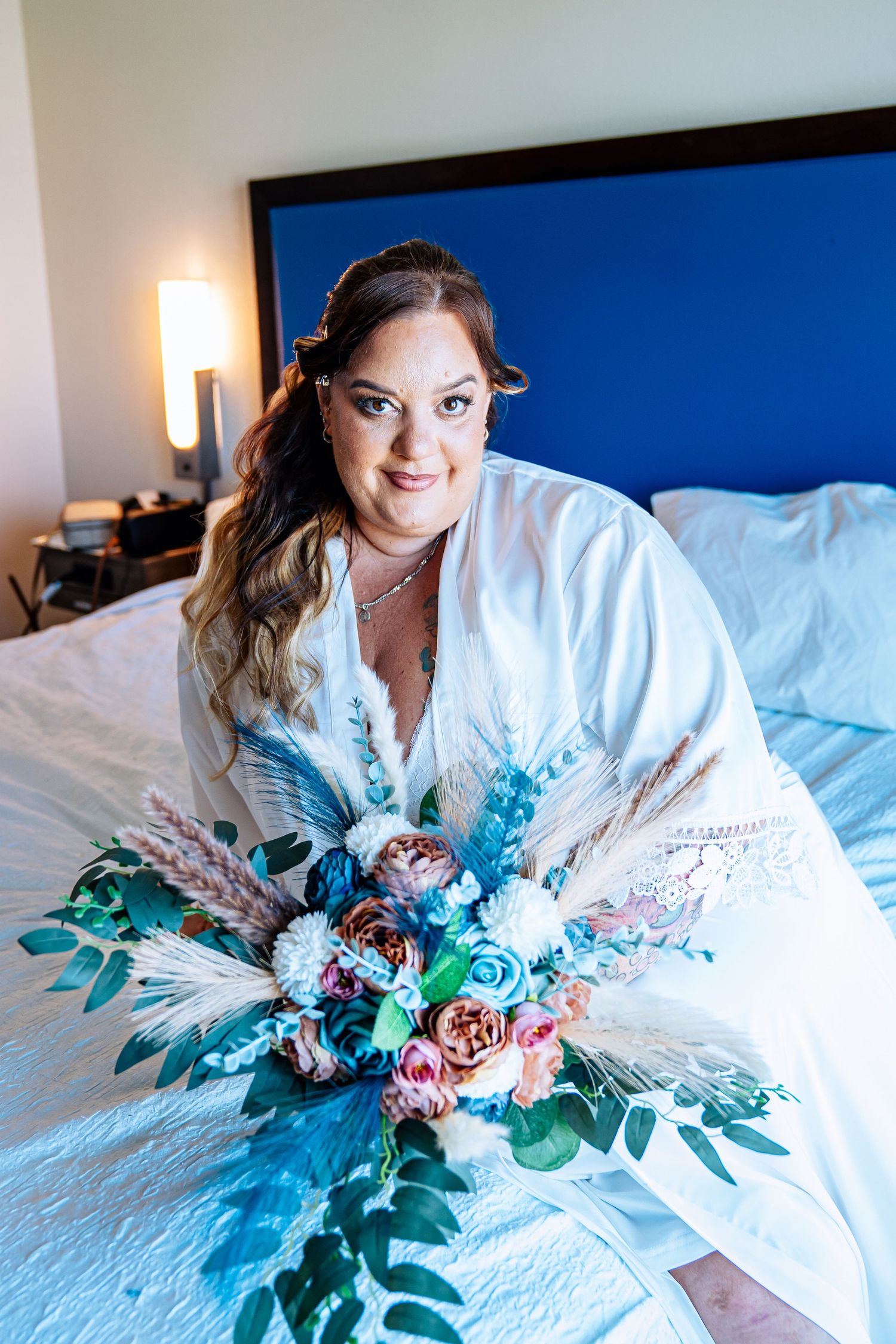 Bride in white robe holding a boho bouquet of teal and pink flowers on a hotel bed before her wedding.