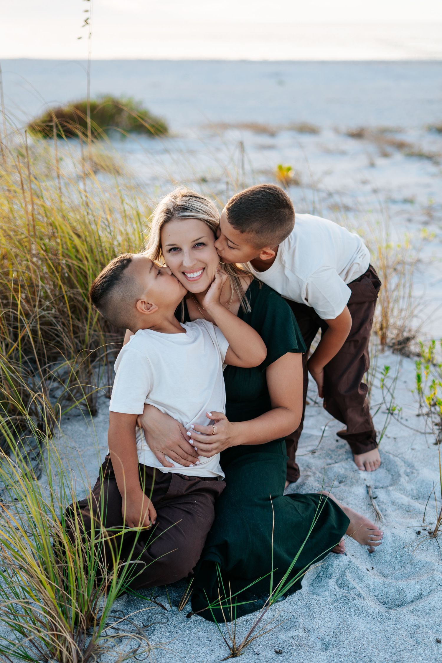 A family shares tender moments together while sitting on white sand beach near sea grass.