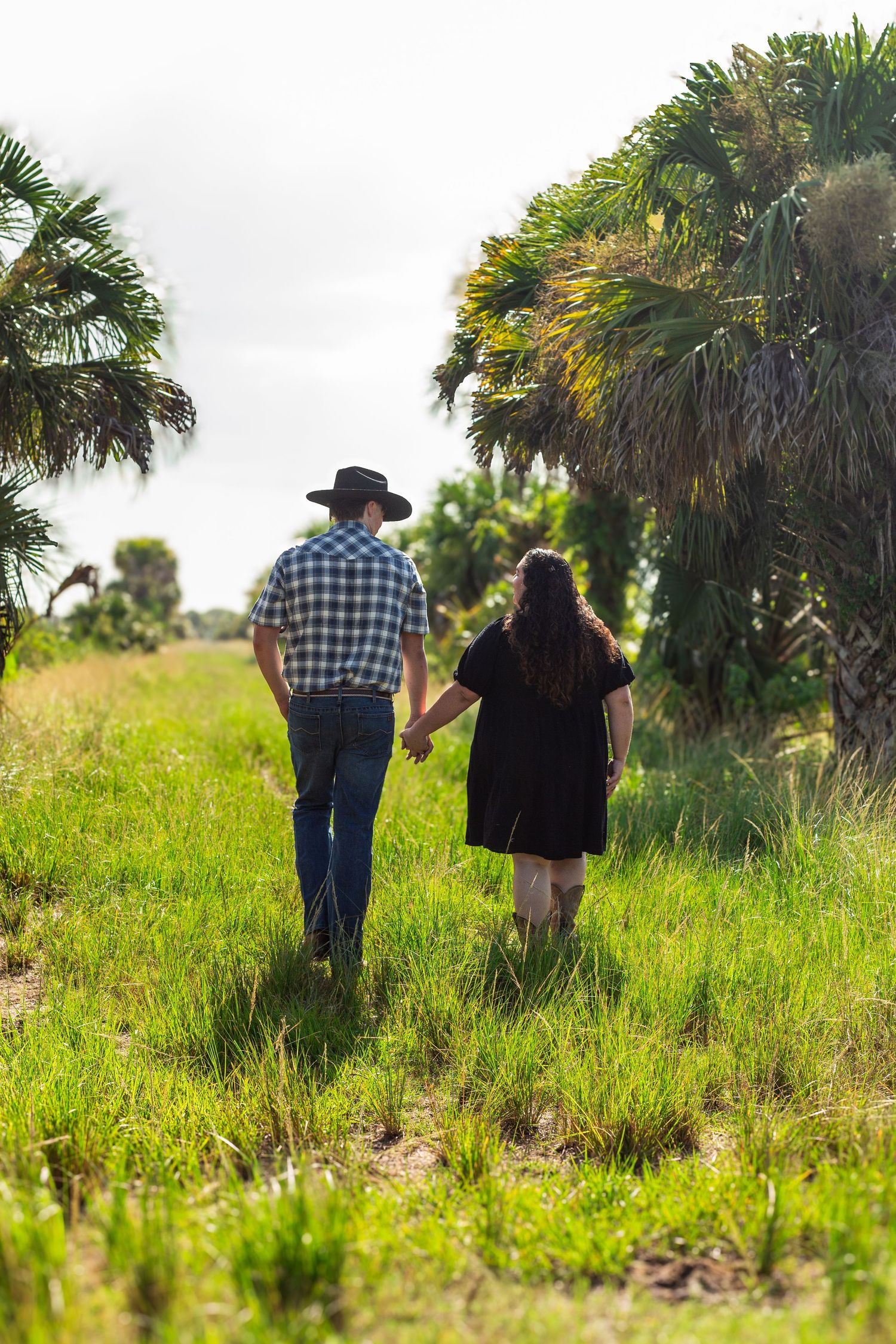 A couple walks hand in hand through tall grass and palm trees on an overcast day.