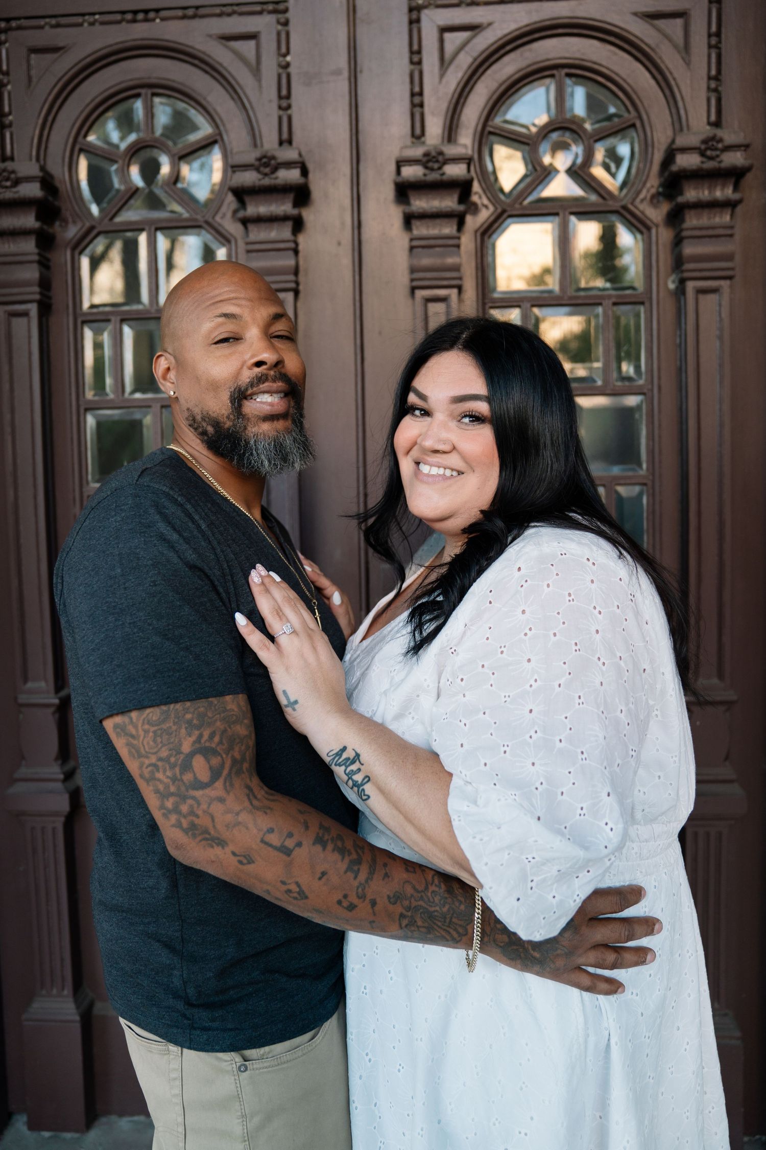 Couple embracing and smiling in front of ornate wooden doors, man with tattoos in gray shirt, woman in white dress.
