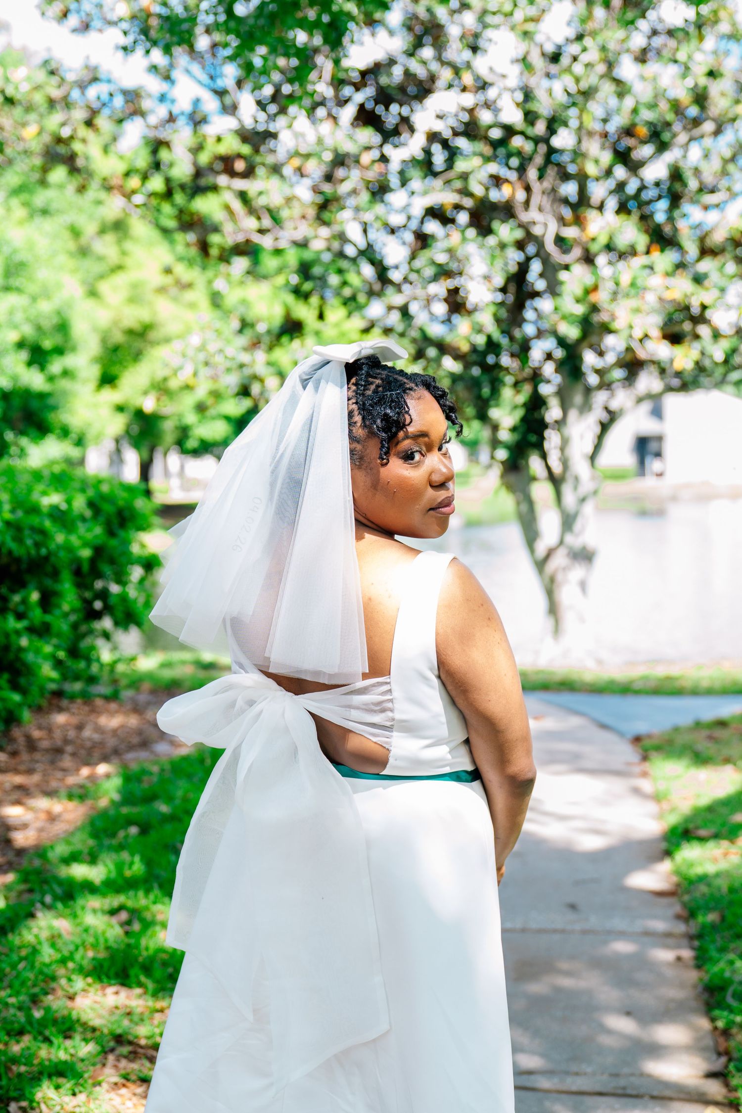 Bride in white gown and veil poses outdoors on a garden path, looking over her shoulder gracefully.