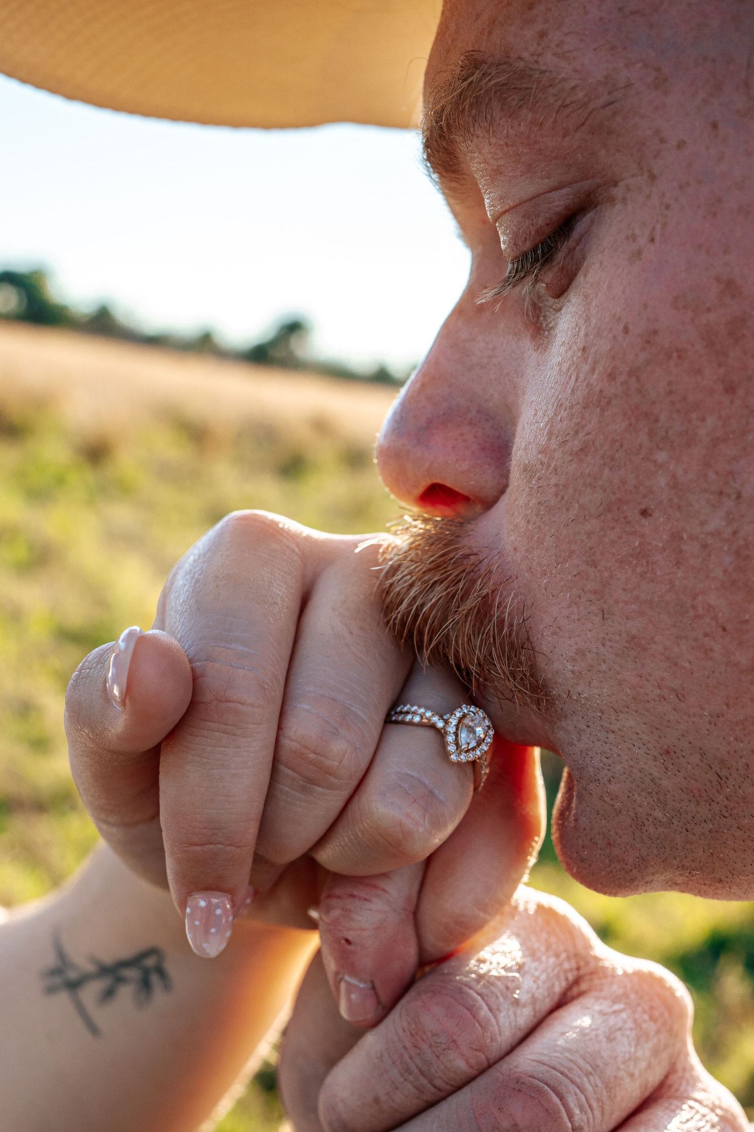 Close-up of a man kissing a woman's hand adorned with a diamond halo engagement ring in an outdoor setting.