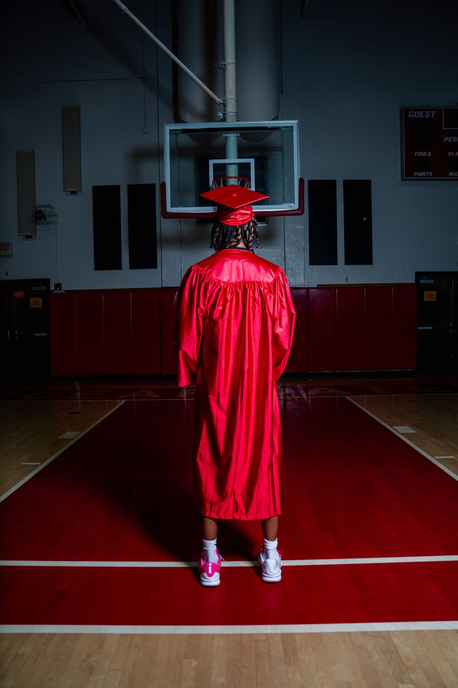 Graduate in red cap and gown stands on basketball court, viewed from behind, in school gymnasium.