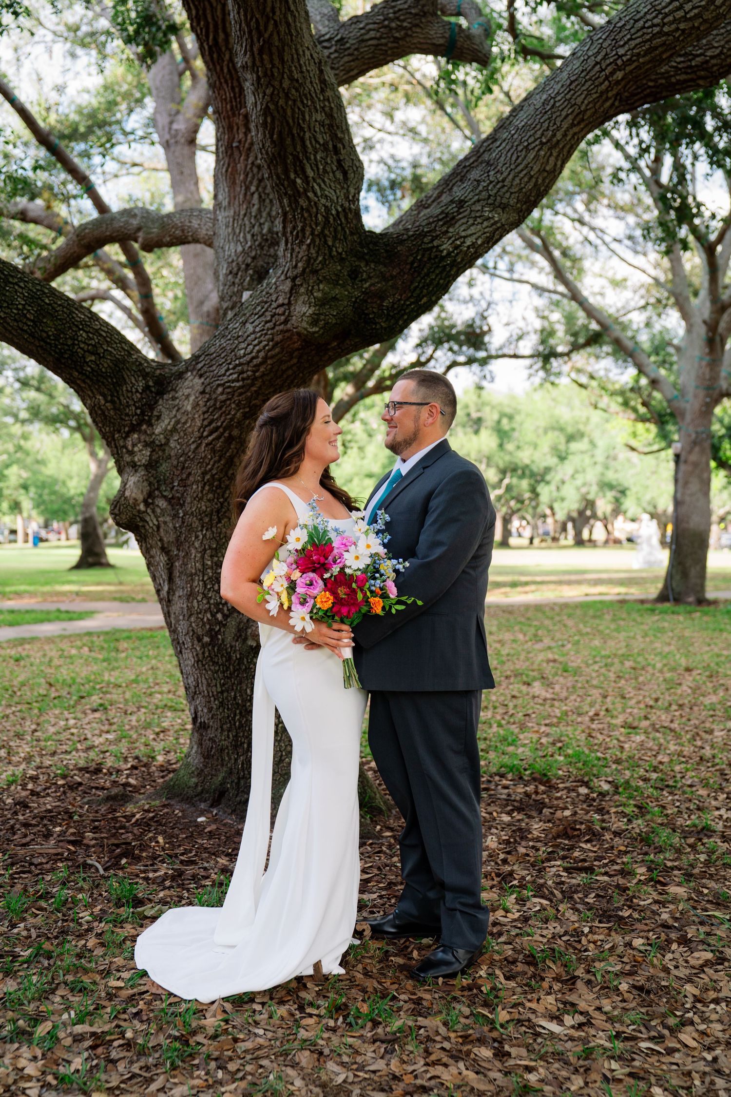 Bride and groom stand beneath a large oak tree in a park, bride holding a vibrant colorful bouquet in a white gown.
