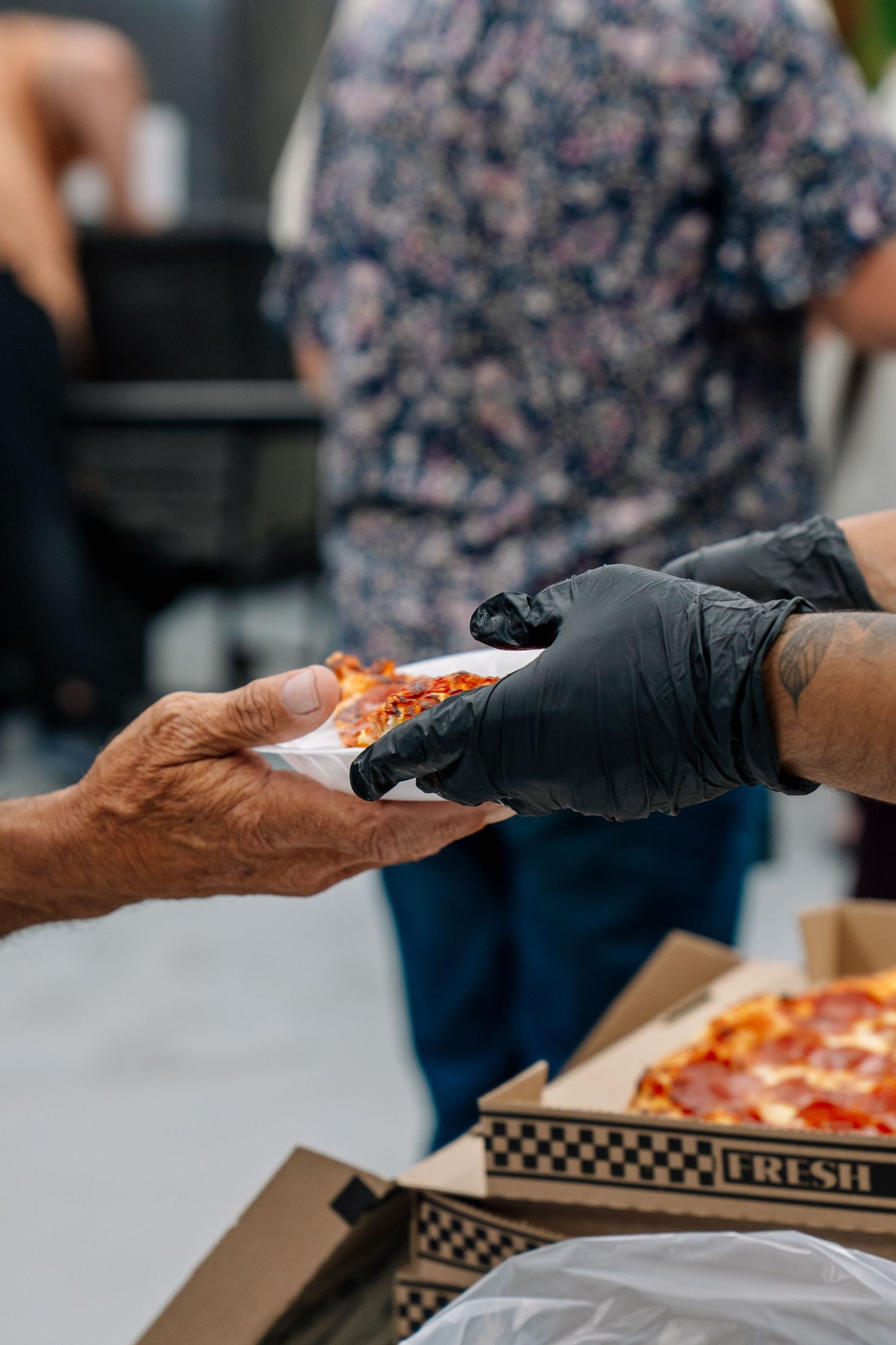 Volunteer in black gloves hands a slice of pizza to an elderly person at an outdoor food drive.