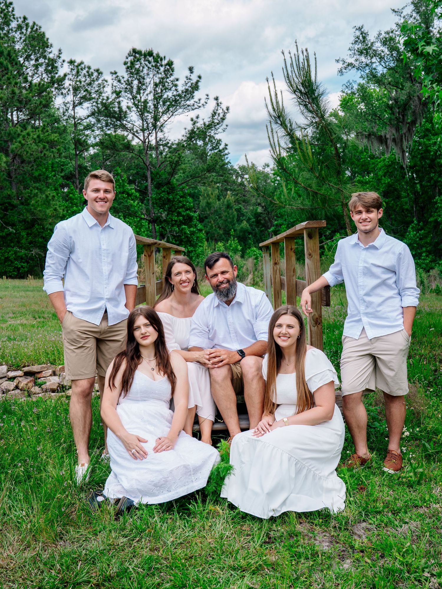 Family of six posing outdoors in coordinated white and khaki outfits on a green lawn with wooden bridge backdrop.