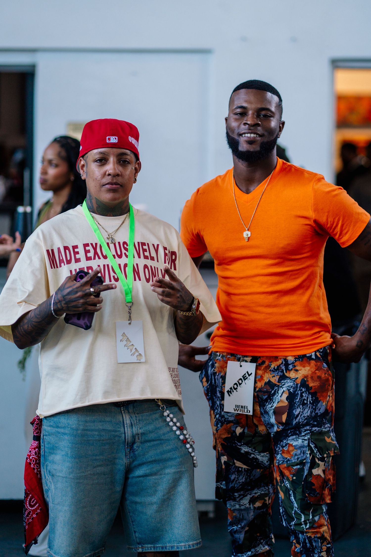 Two men posing together, one in a cream graphic tee and red cap, the other in an orange shirt and floral pants.