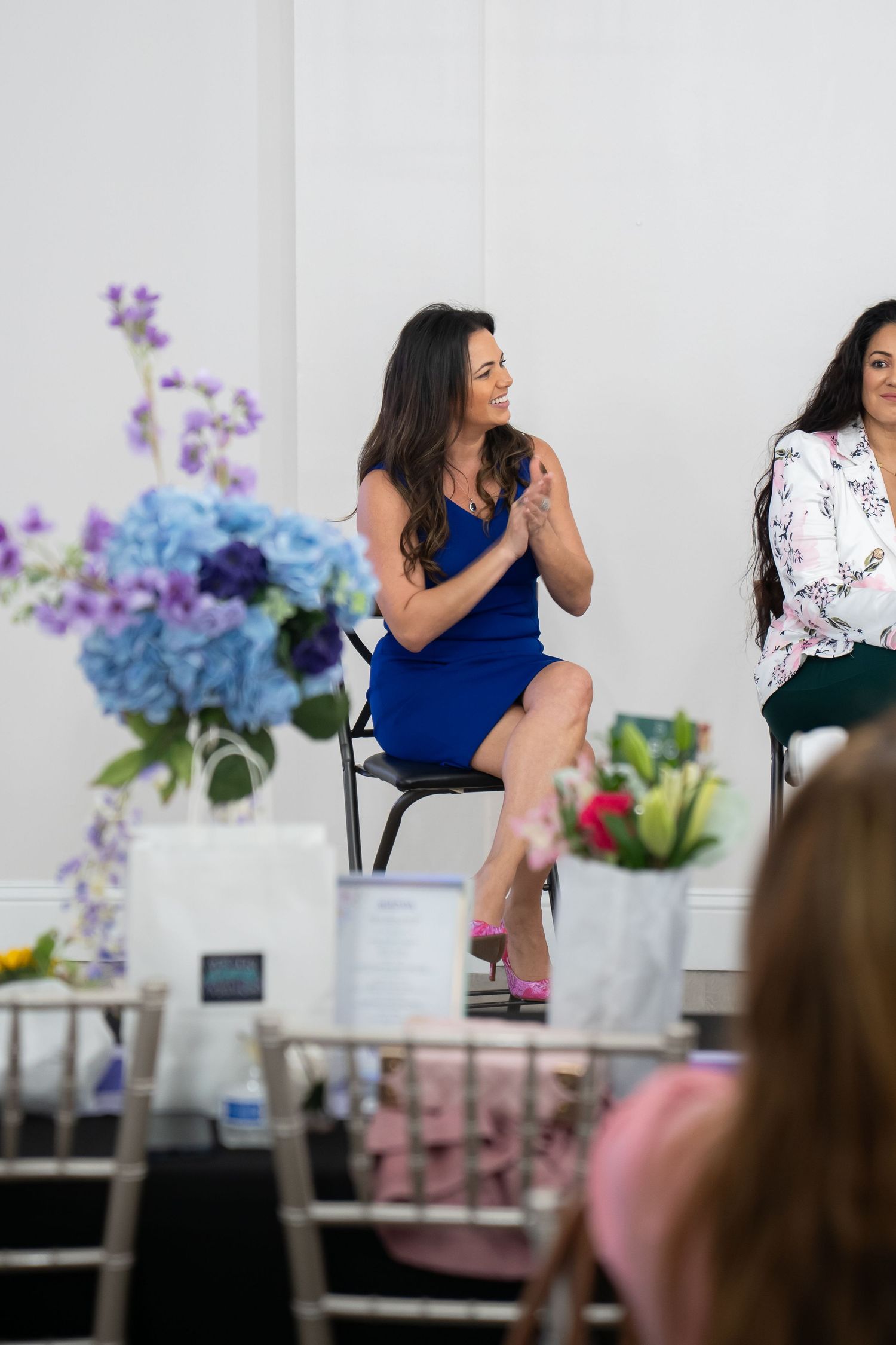 Woman in blue dress speaking at floral event, seated on stool with purple hydrangeas and white flowers in background.
