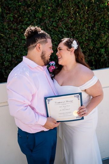 A couple in wedding attire share intimate moments while holding a marriage certificate against a hedge backdrop.
