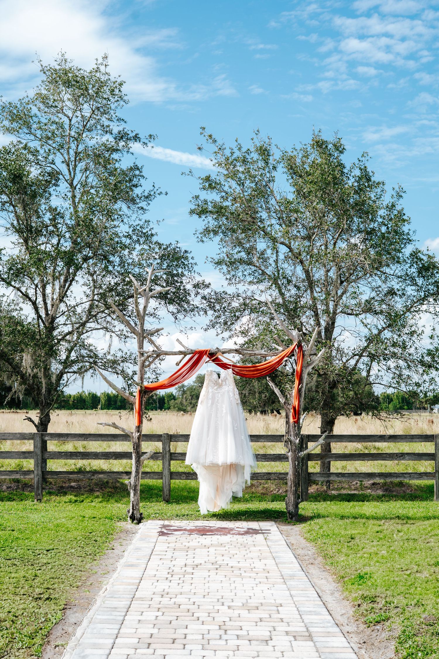 White wedding dress displayed outdoors between trees with red ribbons on a stone pathway.