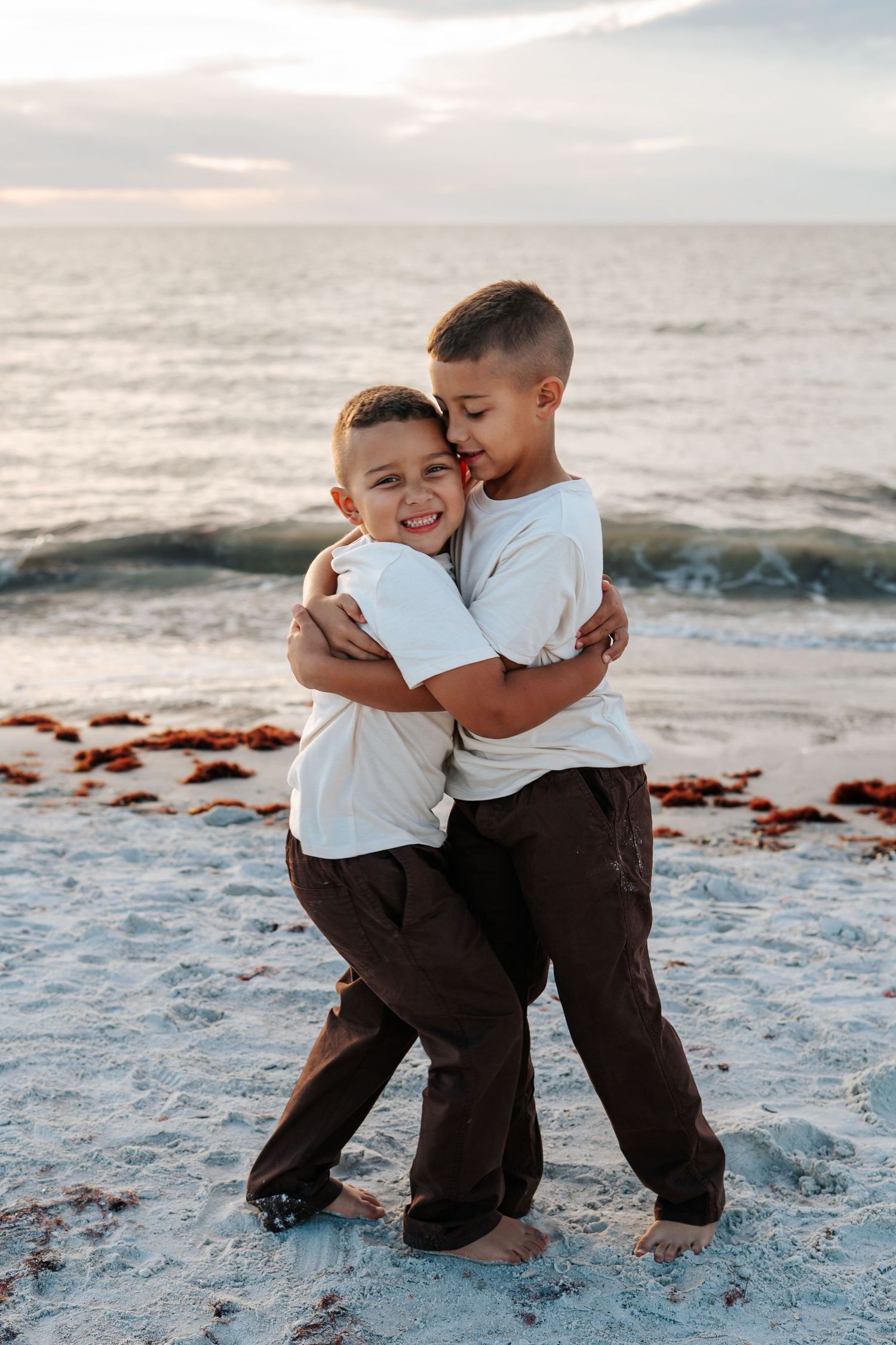 Two young boys in matching outfits share a warm embrace on a sandy beach.