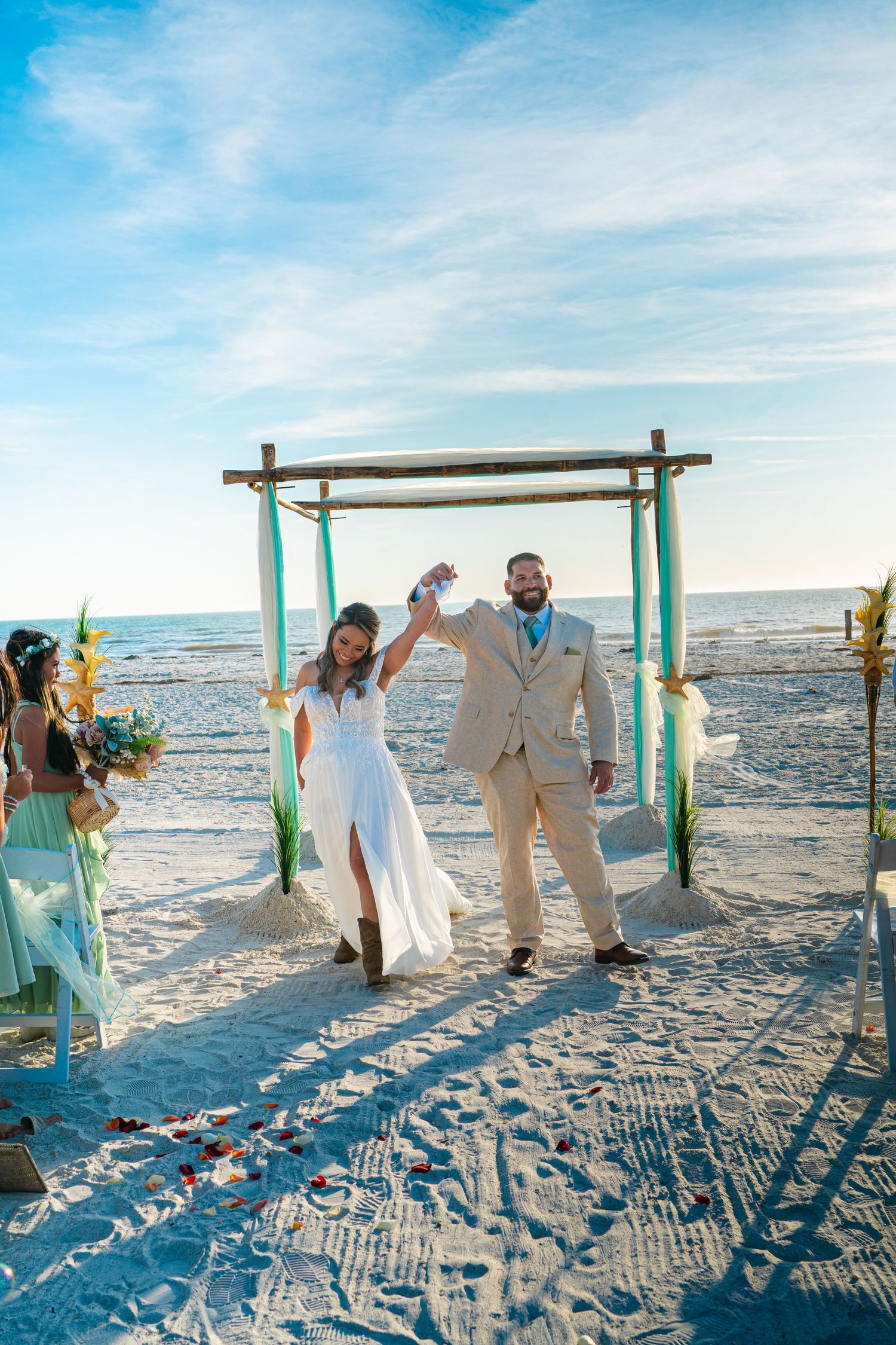 A beach wedding ceremony with a bamboo arch decorated in turquoise fabric as a couple celebrates on white sand by the ocean.