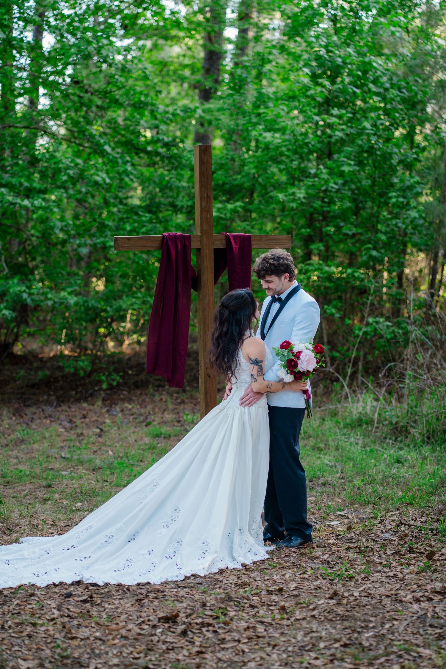 Bride and groom share intimate moment before wooden cross altar in forest setting, bride in white gown with long train.